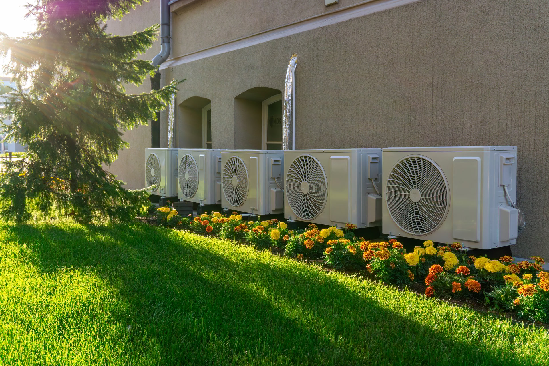 Outdoor air conditioning units mounted on building wall, surrounded by flower bed with yellow and orange flowers, green lawn, sunlight, modern installation in outdoor space.