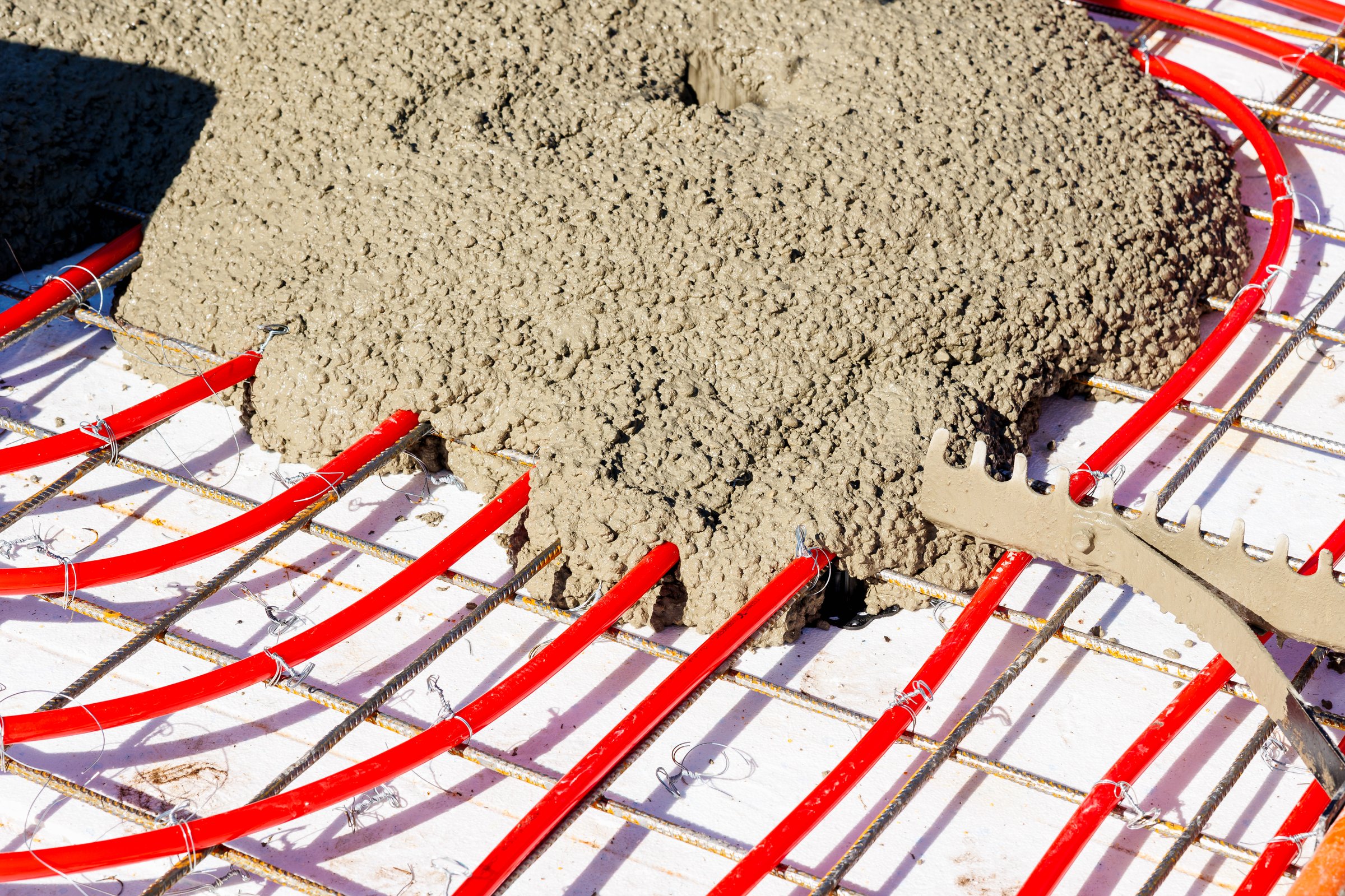 Red underfloor heating pipes attached to a steel mesh grid being covered with freshly poured concrete on a construction site. High quality photo
