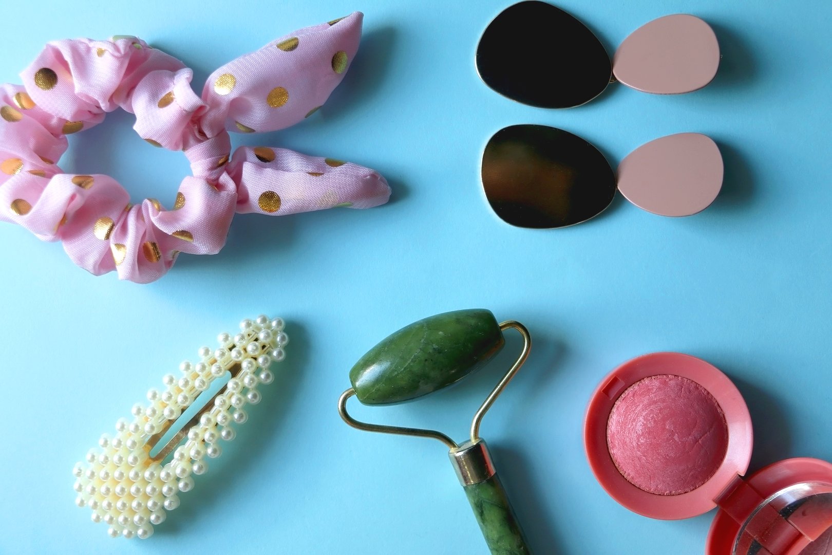 Jade roller, pink blush, fashionable earrigns, pink scrunchie and pearl barrette on bright blue background. Flat lay.
