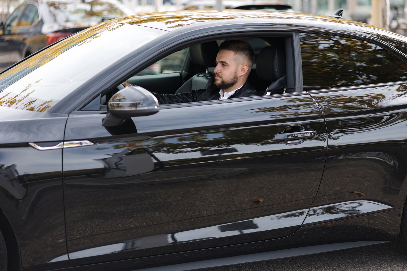 Success in motion. Handsome young man in leather jacket and sunglasses driving a car. Side view from outside . High quality photo