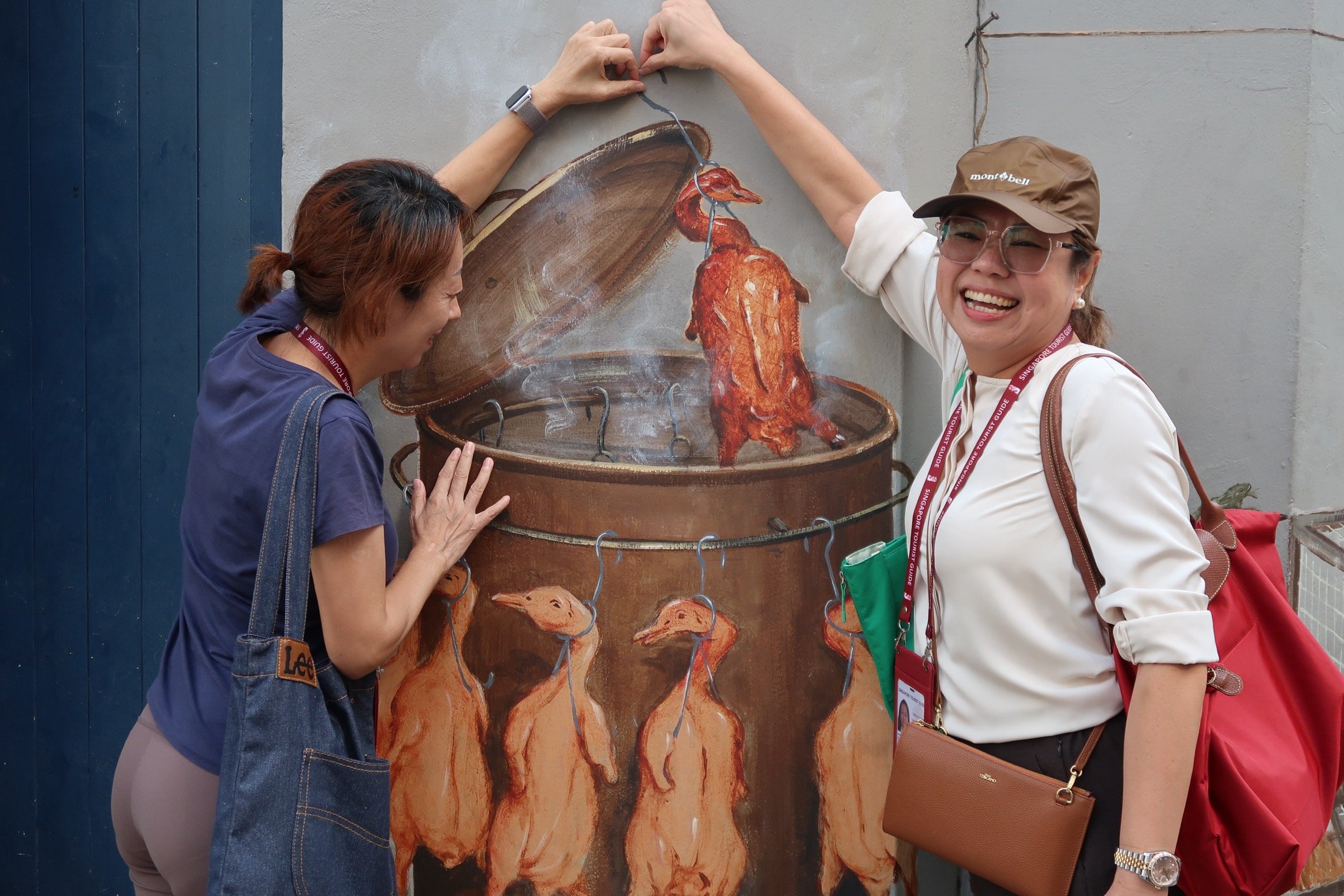 Two women interact with a mural of roast ducks in a barrel, one pretending to close the lid and the other smiling.