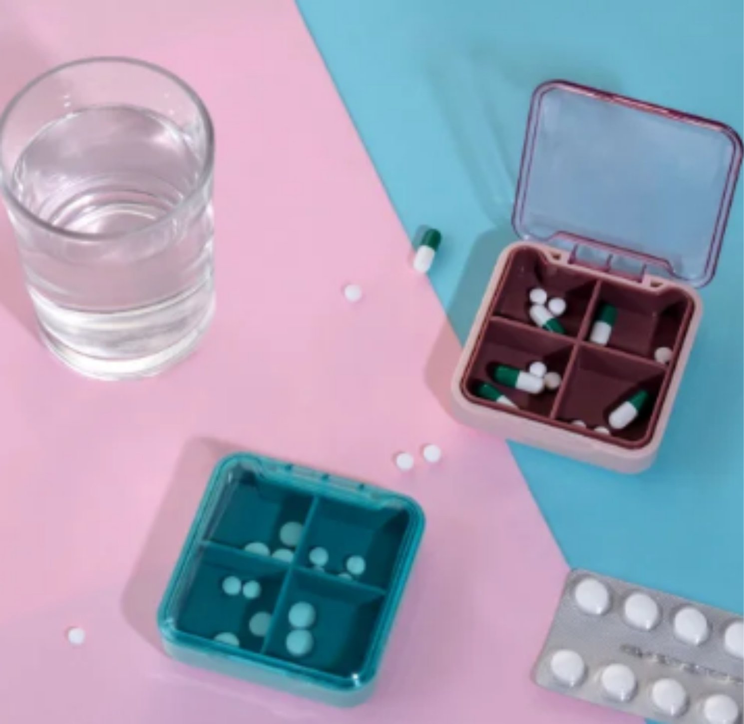 Two pill organizers, a blister pack, loose pills, and a glass of water on pink and blue background.