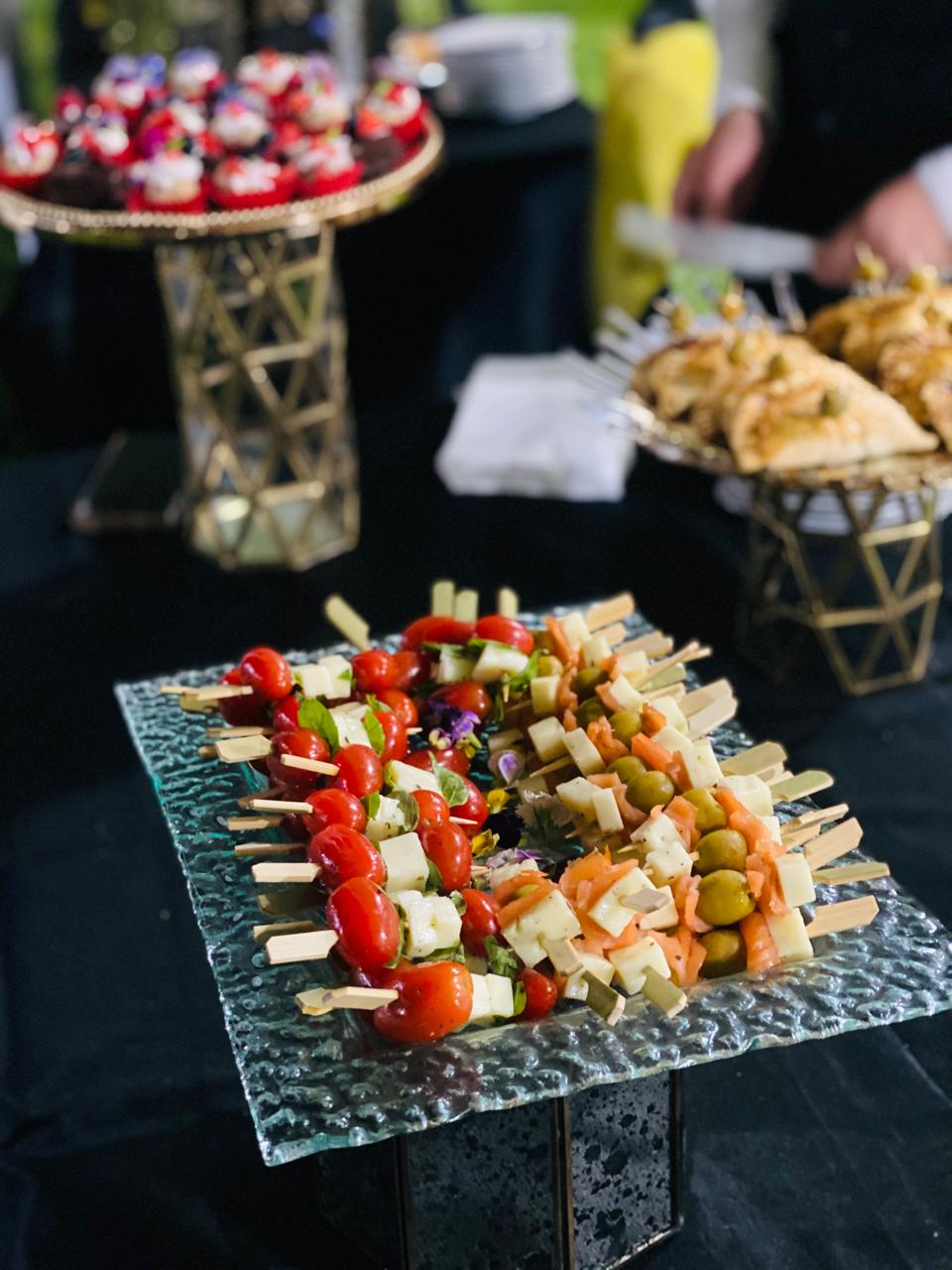 Assorted skewers of vegetables and cheese on a glass platter with pastries and cupcakes in the background at a buffet.