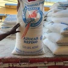 A person holding a large sack of refined sugar labeled 125 lbs, stacked on a platform with other similar bags.