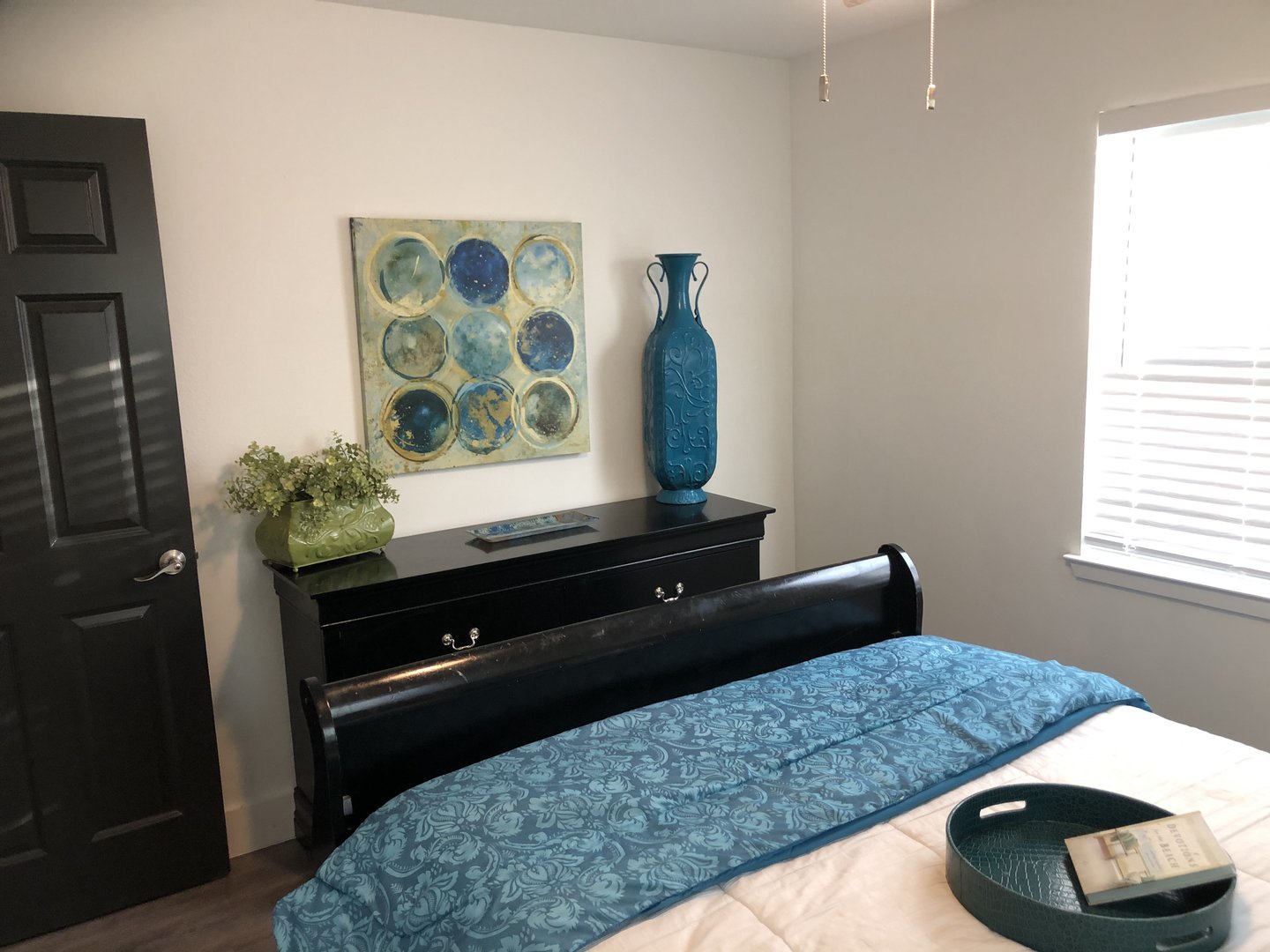 Bedroom with a blue-themed decor, including a vase, bedspread, and artwork, against a neutral wall with a black dresser.