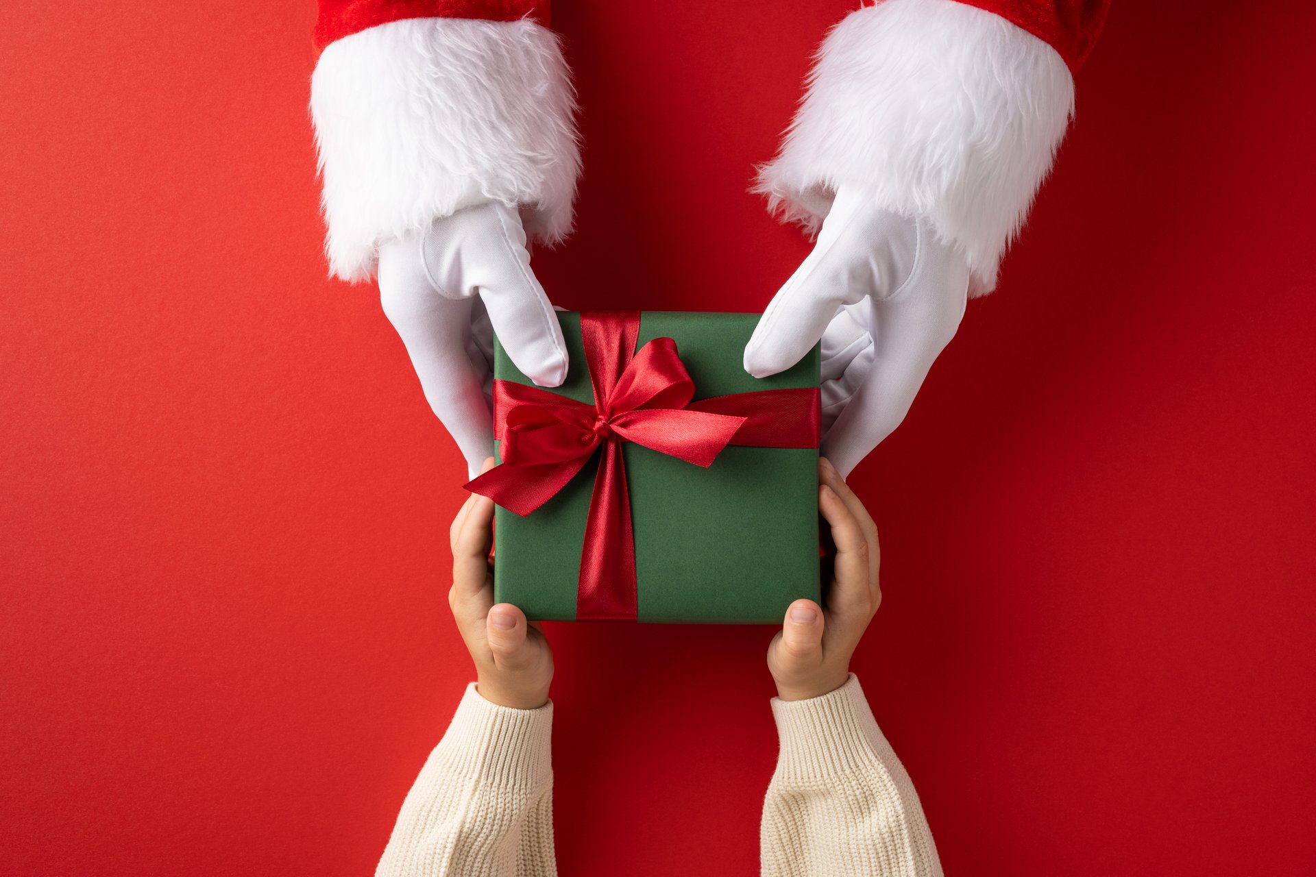 A pair of hands in Santa gloves gives a gift wrapped in a green box to a child's hands, featuring a vibrant red background symbolizing holiday cheer