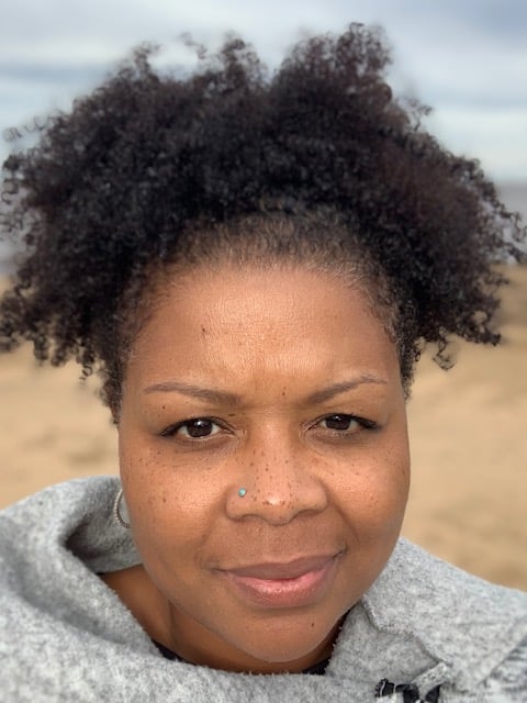 Person outdoors with curly hair, wearing a gray sweater. The background is a sandy landscape with a cloudy sky.