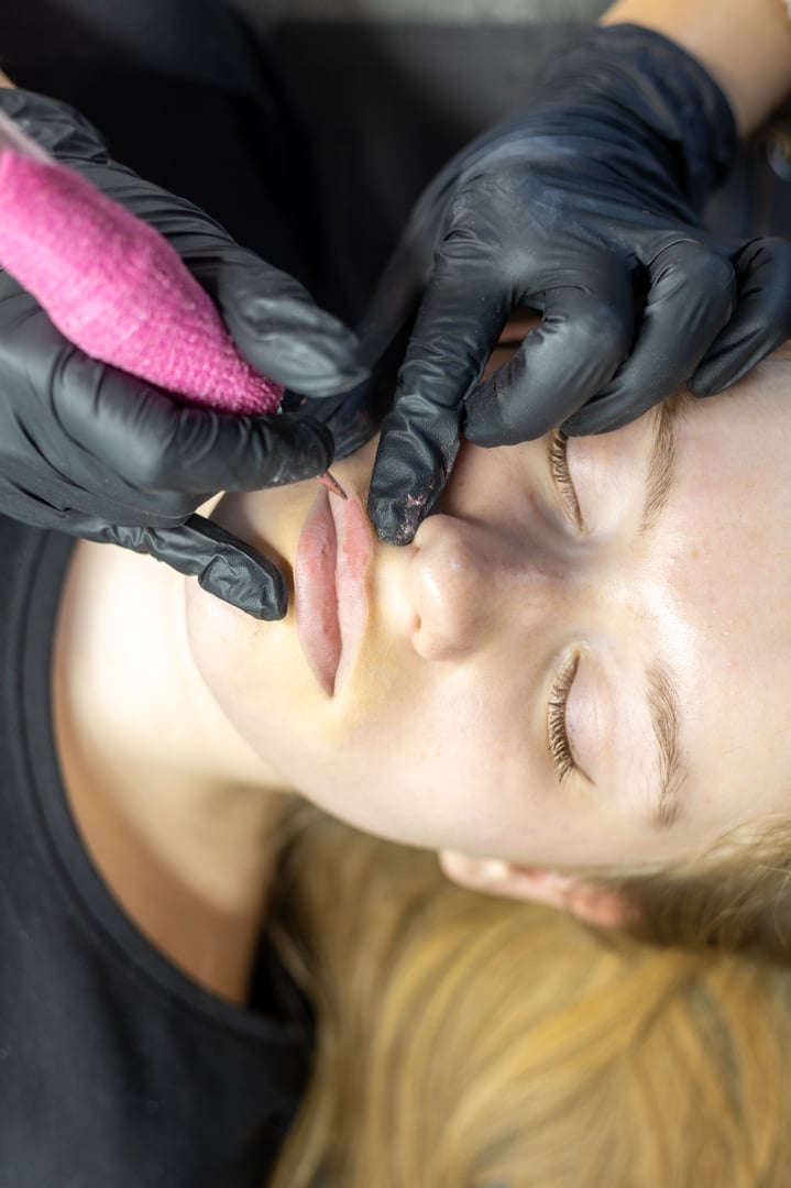 A beauty expert meticulously applies permanent makeup to a client's lips using a precision tool. The client is relaxed as the artist focuses on enhancing her natural features in a salon.