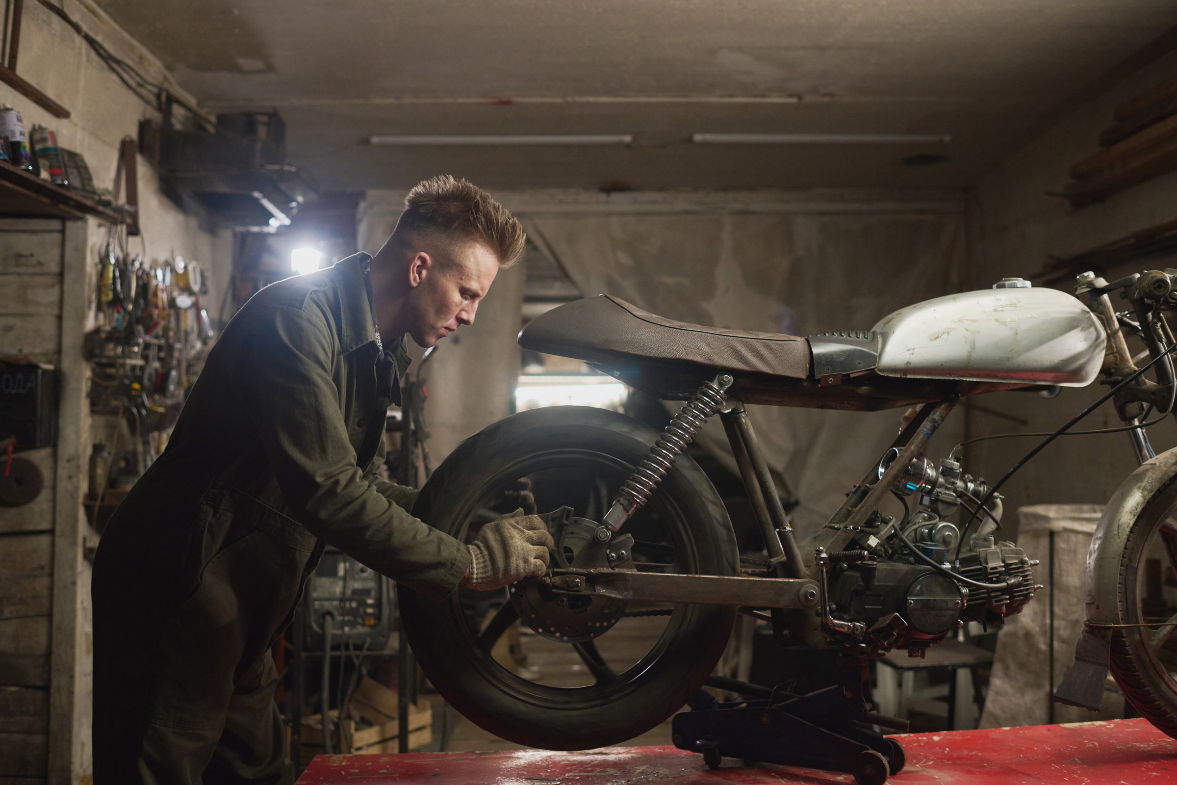 Young adult Caucasian man repairing custom motorcycle in workshop, focusing on adjusting rear wheel and suspension components, surrounded by tools and mechanical equipment