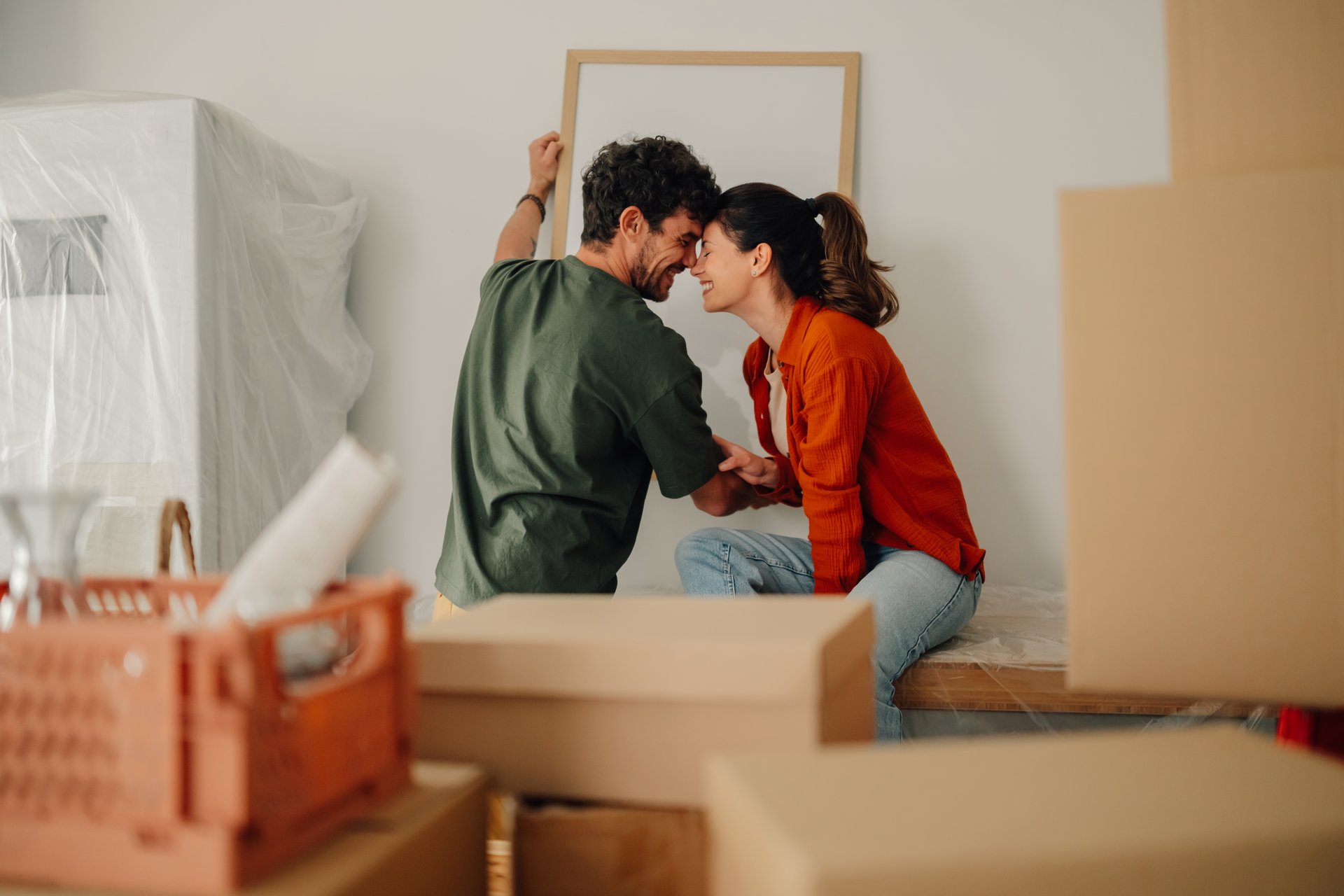 Young couple hanging a picture frame on the wall in their new apartment, surrounded by cardboard boxes, smiling and touching foreheads