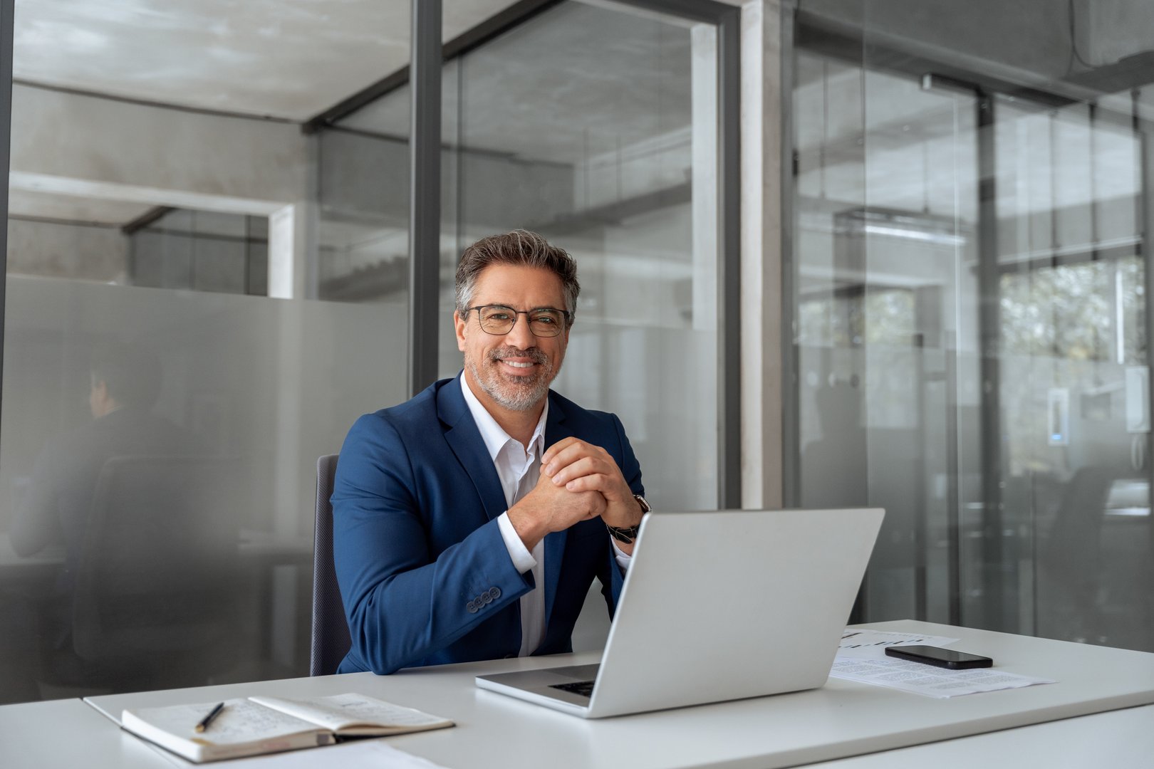 Portrait of mature Indian or Latin hispanic business man ceo manager using laptop computer in modern office. Middle eastern smiling senior businessman entrepreneur working on pc, looking at camera