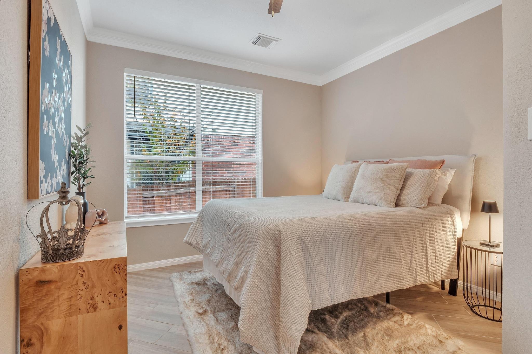 Cozy bedroom with a double bed, soft pillows, a wooden dresser, and a large window with blinds, allowing natural light in.
