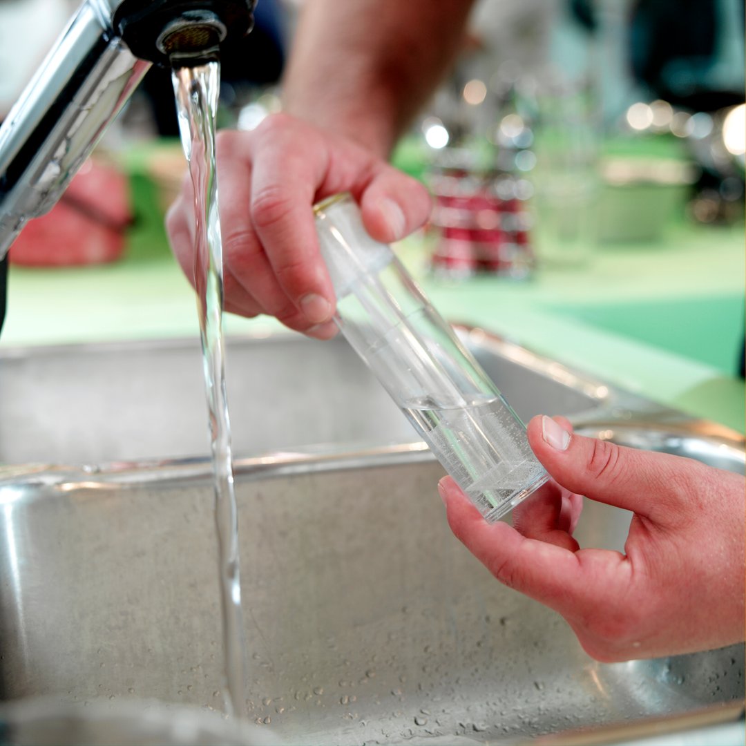 Person rinsing a clear bottle under a kitchen tap with running water inside a stainless steel sink.