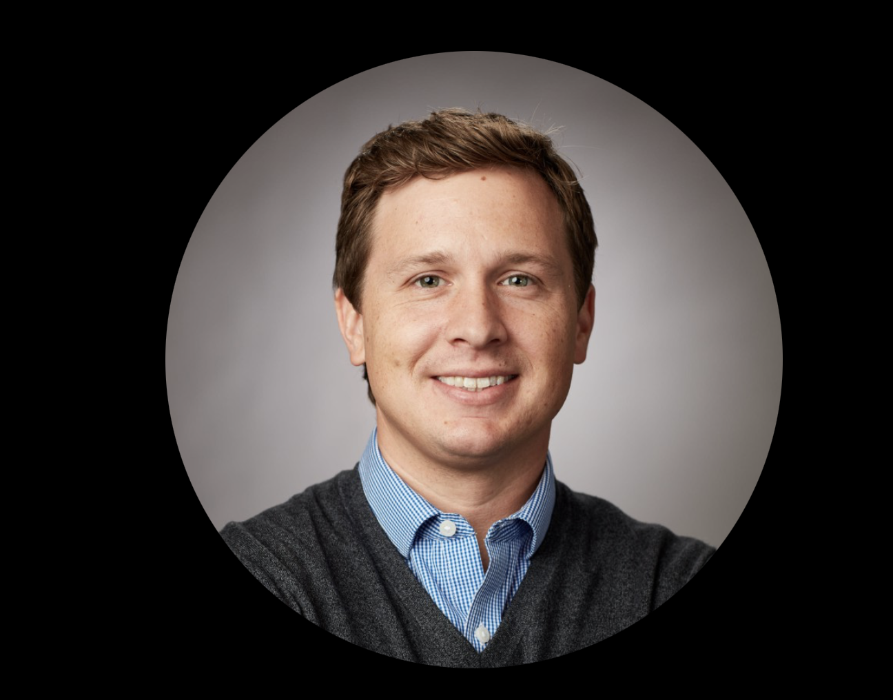 A man with short brown hair wearing a blue shirt and dark sweater, smiling in a circular portrait on a gray background.