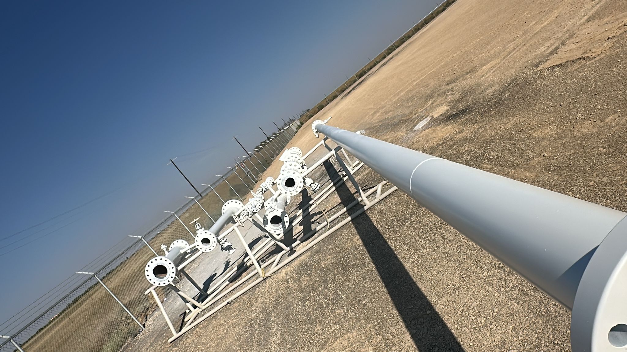 Industrial pipes and fittings arranged on a fenced field under a clear blue sky.