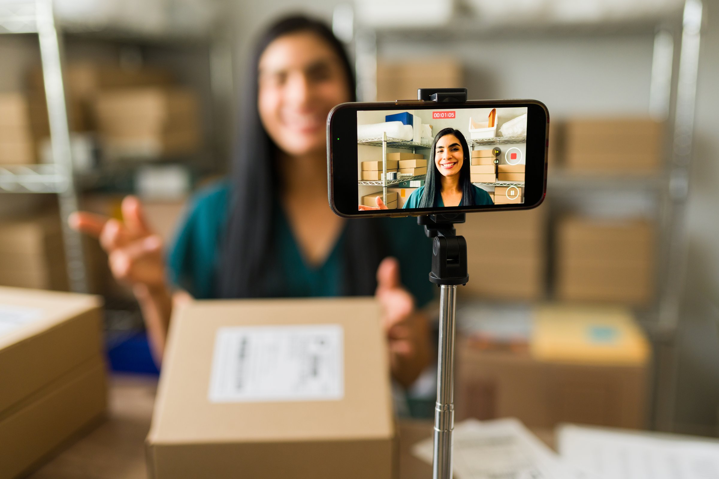 Entrepreneurial latin individual live streaming on social media with a smartphone and a selfie stick while operating in a warehouse