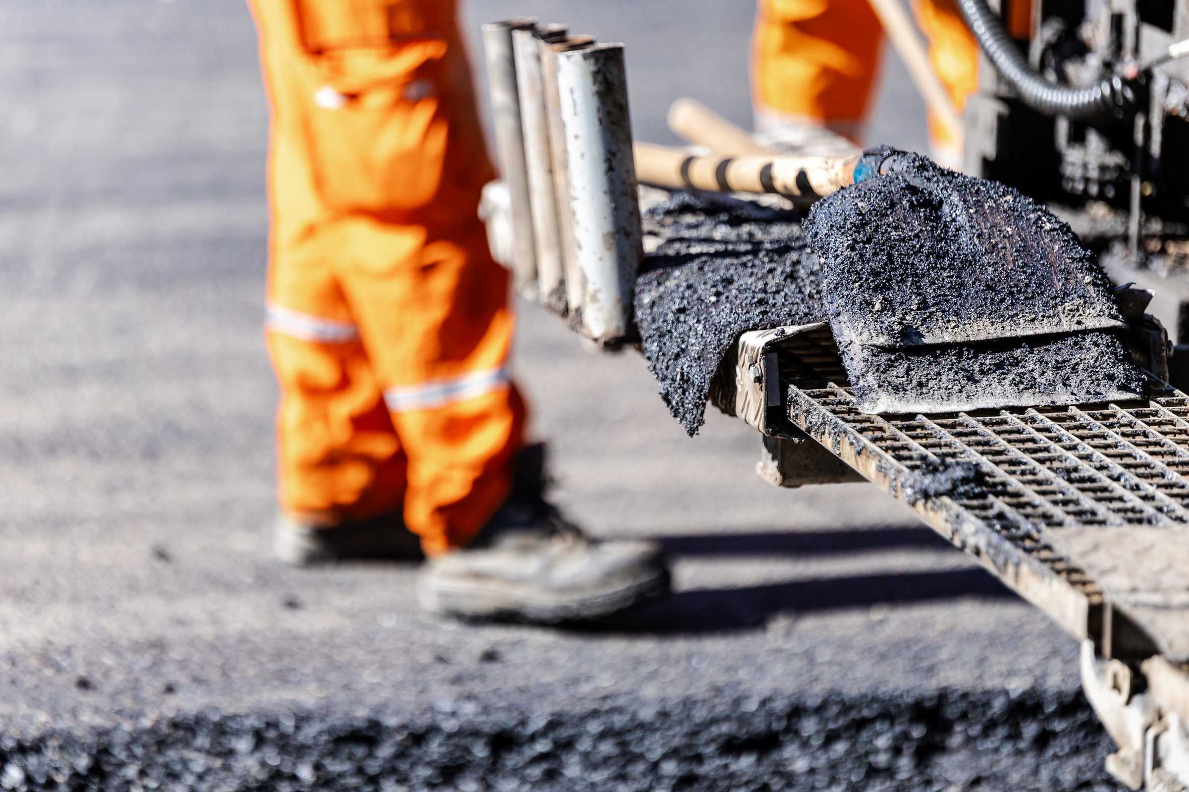 A construction worker in bright orange gear meticulously operates an asphalt paving machine.