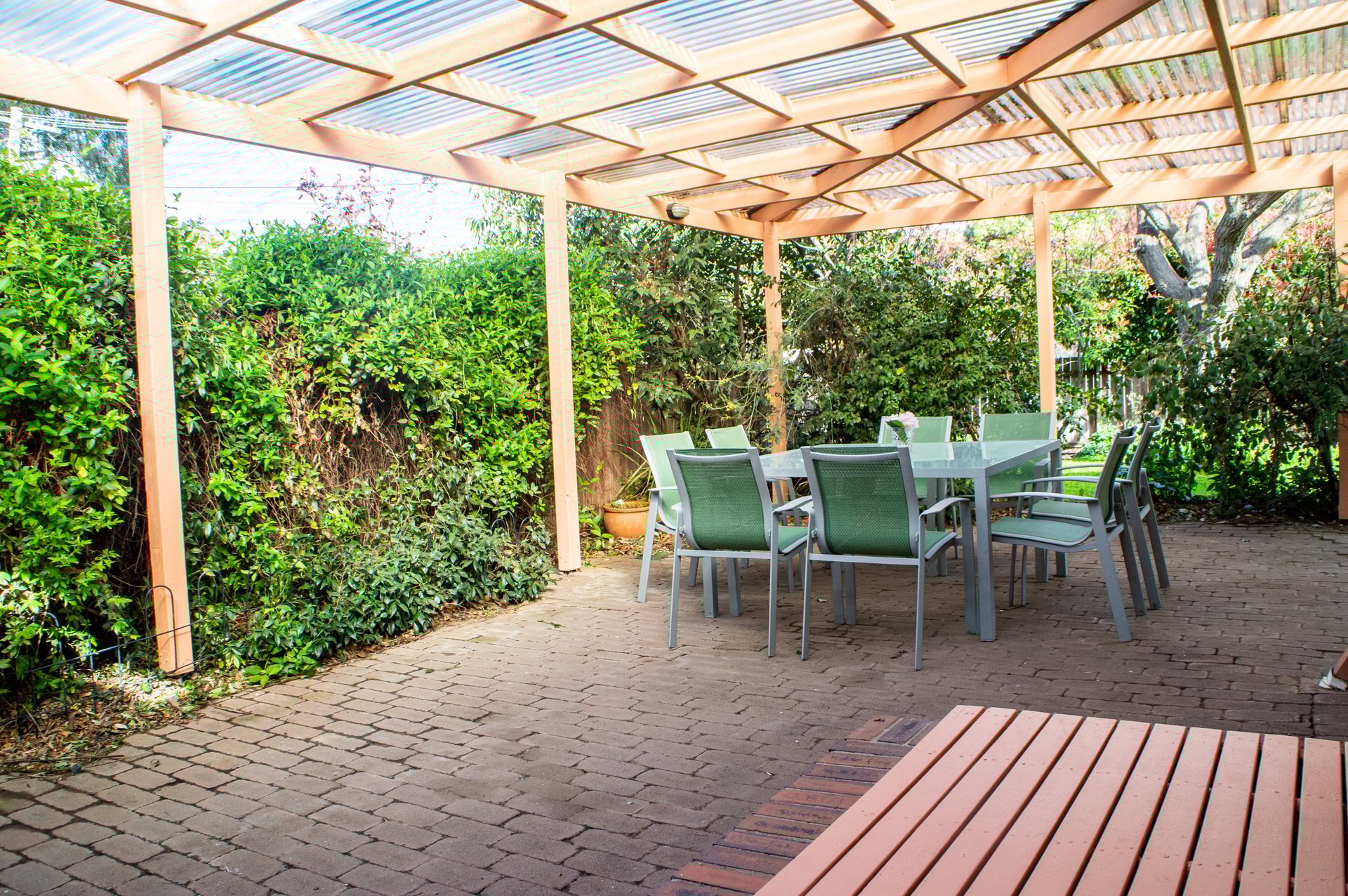 A deck and outdoor table and chairs can be seen under an attractive pergola surrounded by a honeysuckle hedge and other plants.  This makes a spacious covered outdoor area for entertaining.