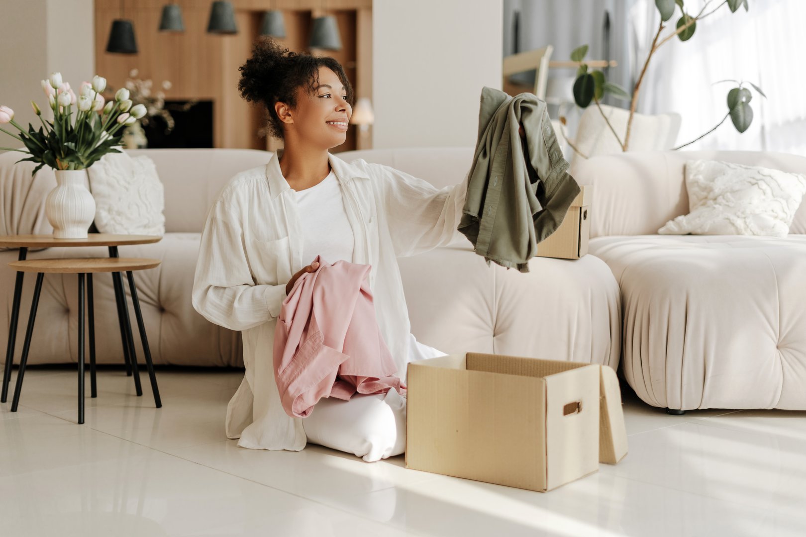 Young woman is smiling while unpacking clothes from a cardboard box in her living room