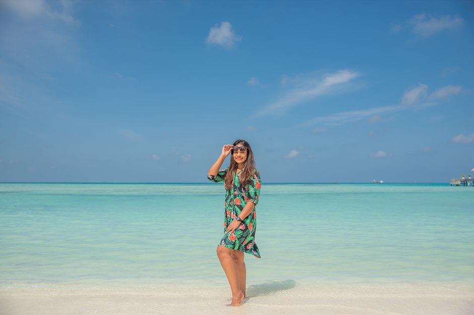 A woman in a floral dress stands on a sandy beach, smiling and adjusting her sunglasses, with clear blue water and sky in the background.