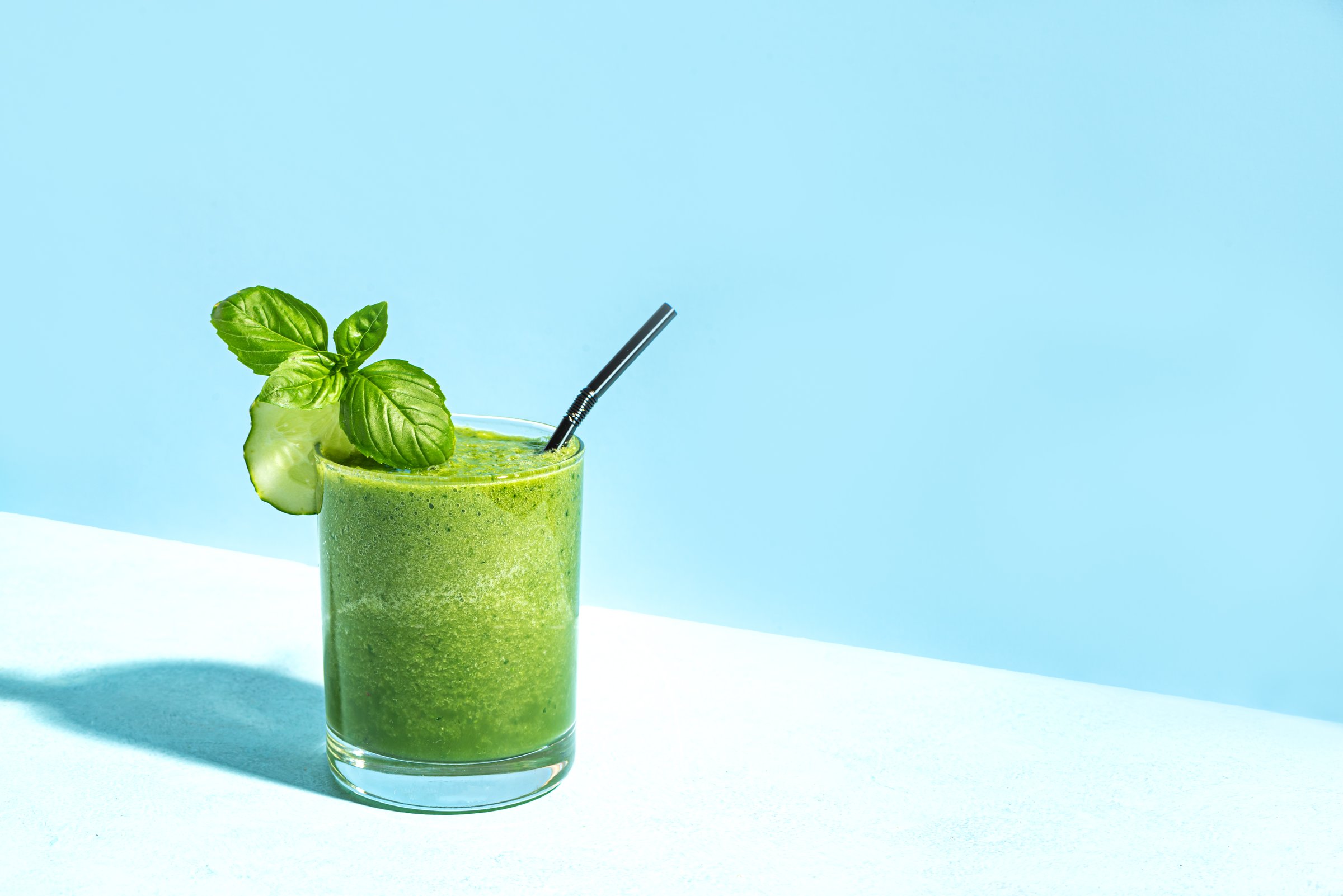 Green smoothie in glass with straw on light blue background, trendy hard light, copy space. Creative advertising concept of detox or clean, healthy food.