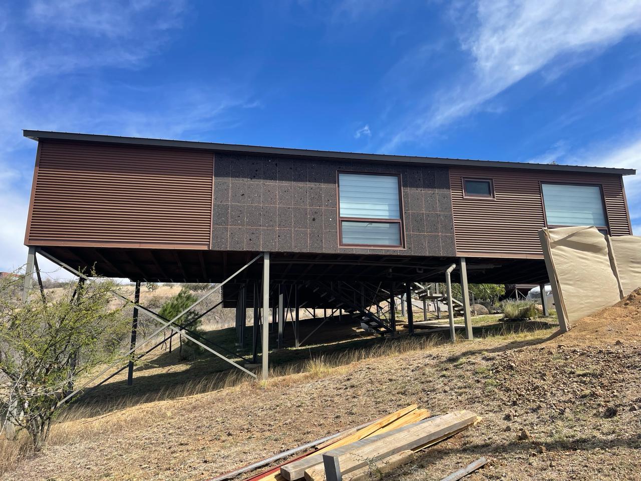Elevated house on stilts with modern design and large windows, set on a hillside under a clear blue sky.