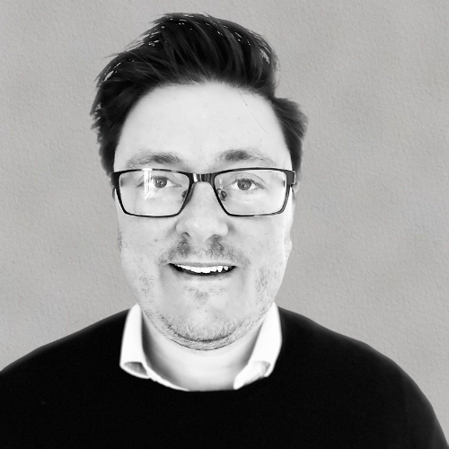 A man with glasses and short dark hair, wearing a black sweater over a white collared shirt, smiling against a plain background.