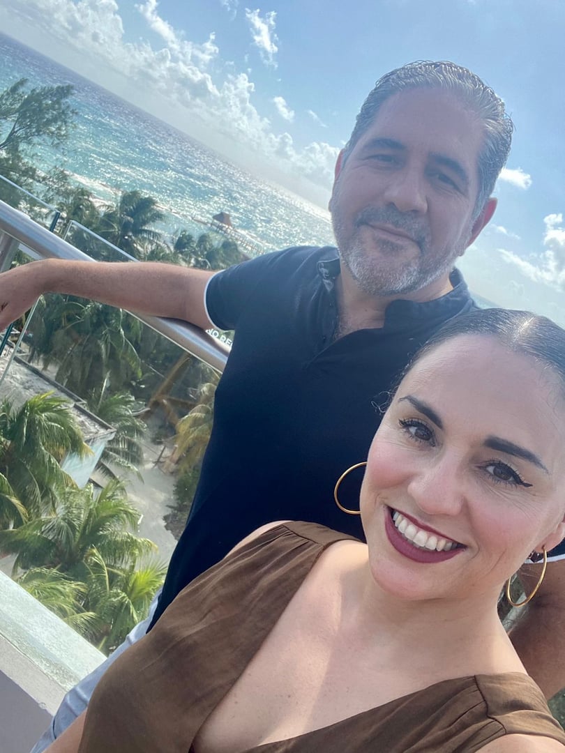 A man and woman smiling on a balcony with a tropical ocean view in the background.