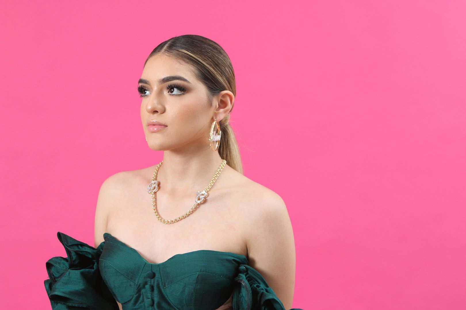 Woman in a green off-shoulder top with gold necklace and earrings, posed against a pink background.
