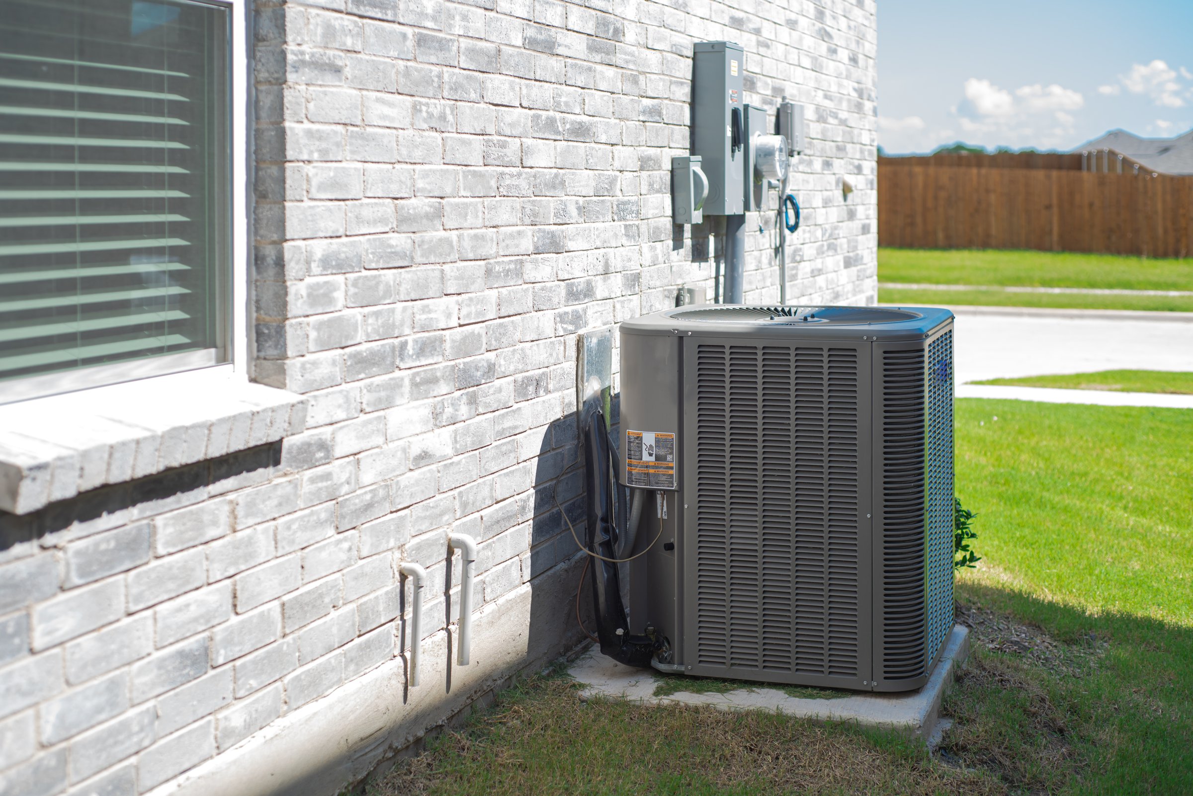 Side windows of new construction home and central air conditioning condenser unit mounted on concrete slab beside brick house wall, utility meters and electrical disconnect, suburbs Dallas, Texas. USA