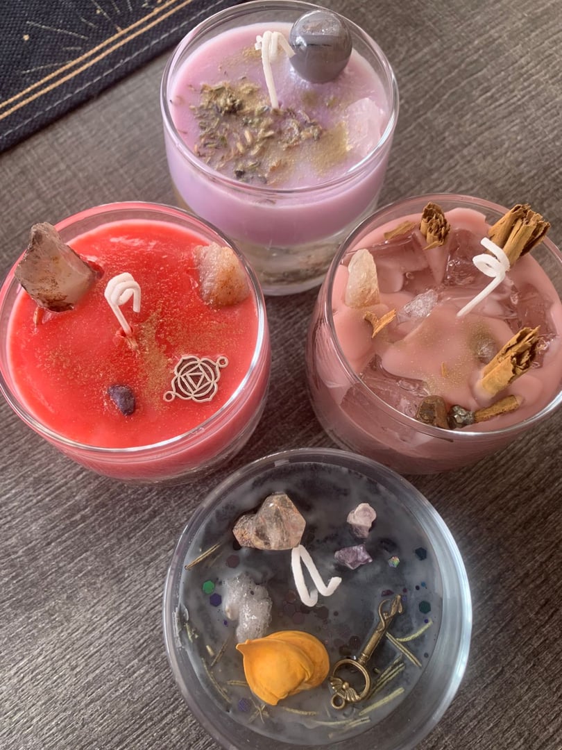 Four decorative candles with crystals, herbs, and embellishments on a table, each in a different color.