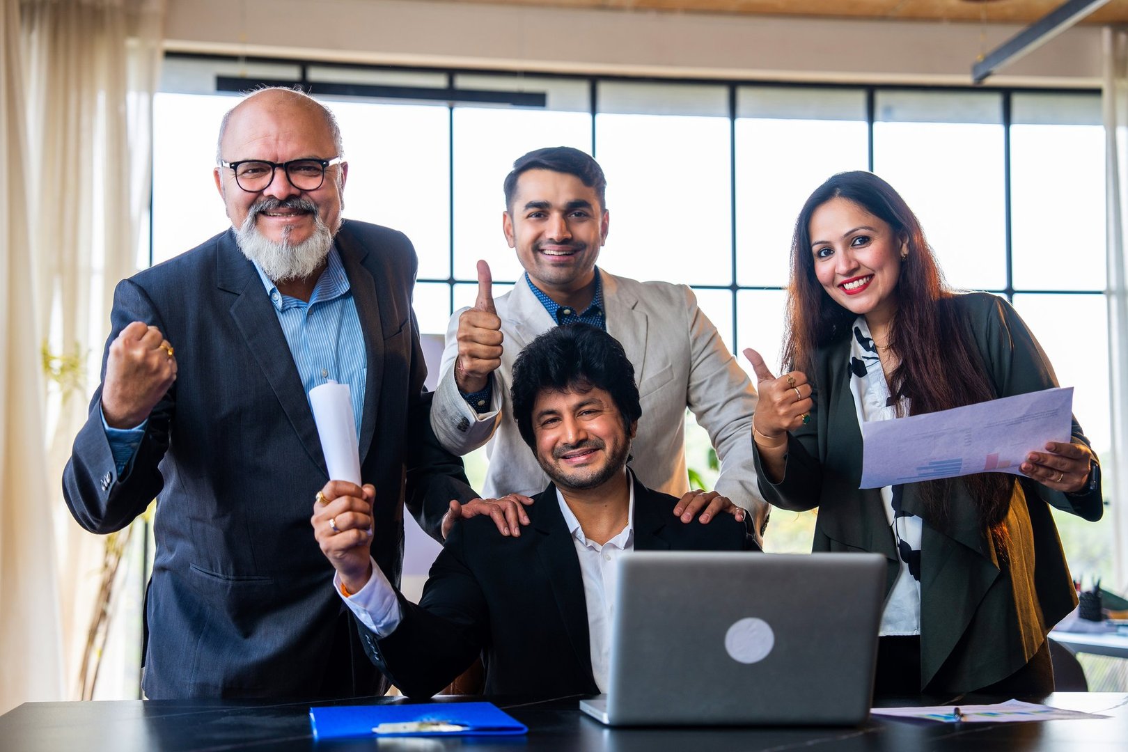 Indian Asian group of confident corporate professionals in formal attire working on laptop and celebrating success with high five and cheerful smiles in office