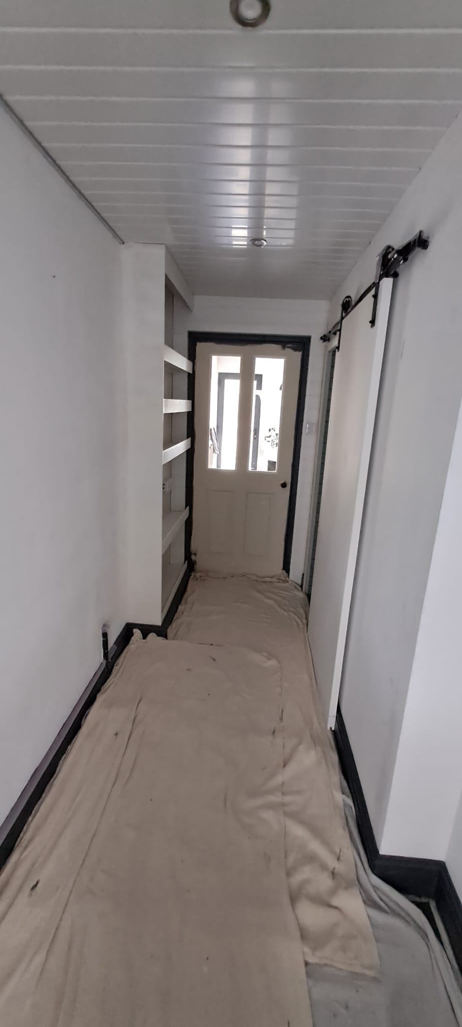 Narrow hallway with white walls, a closed door at the end, and shelves on the left. Drop cloths cover the floor.