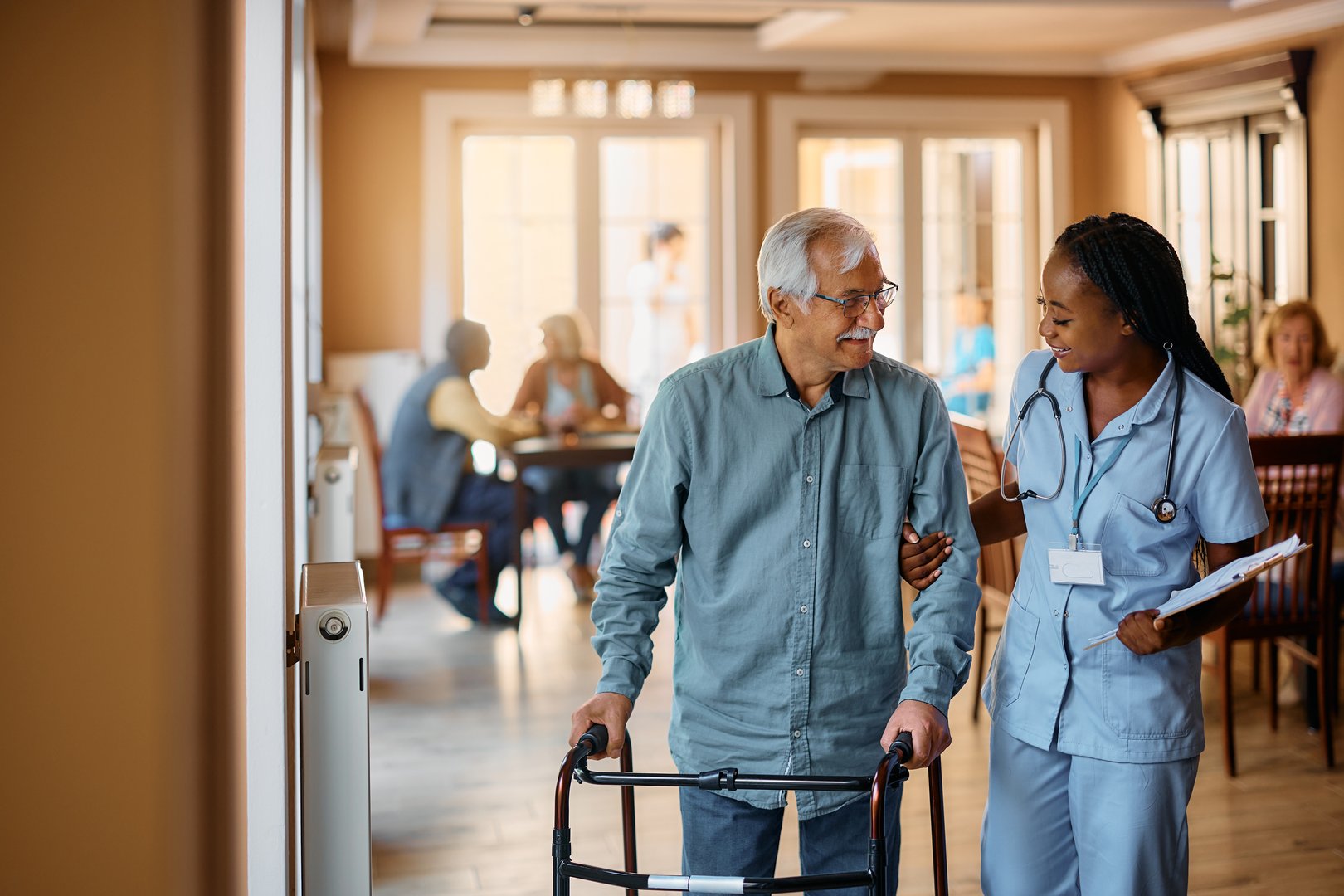 Happy senior man using mobility walker while African American nurse is helping him in nursing home.