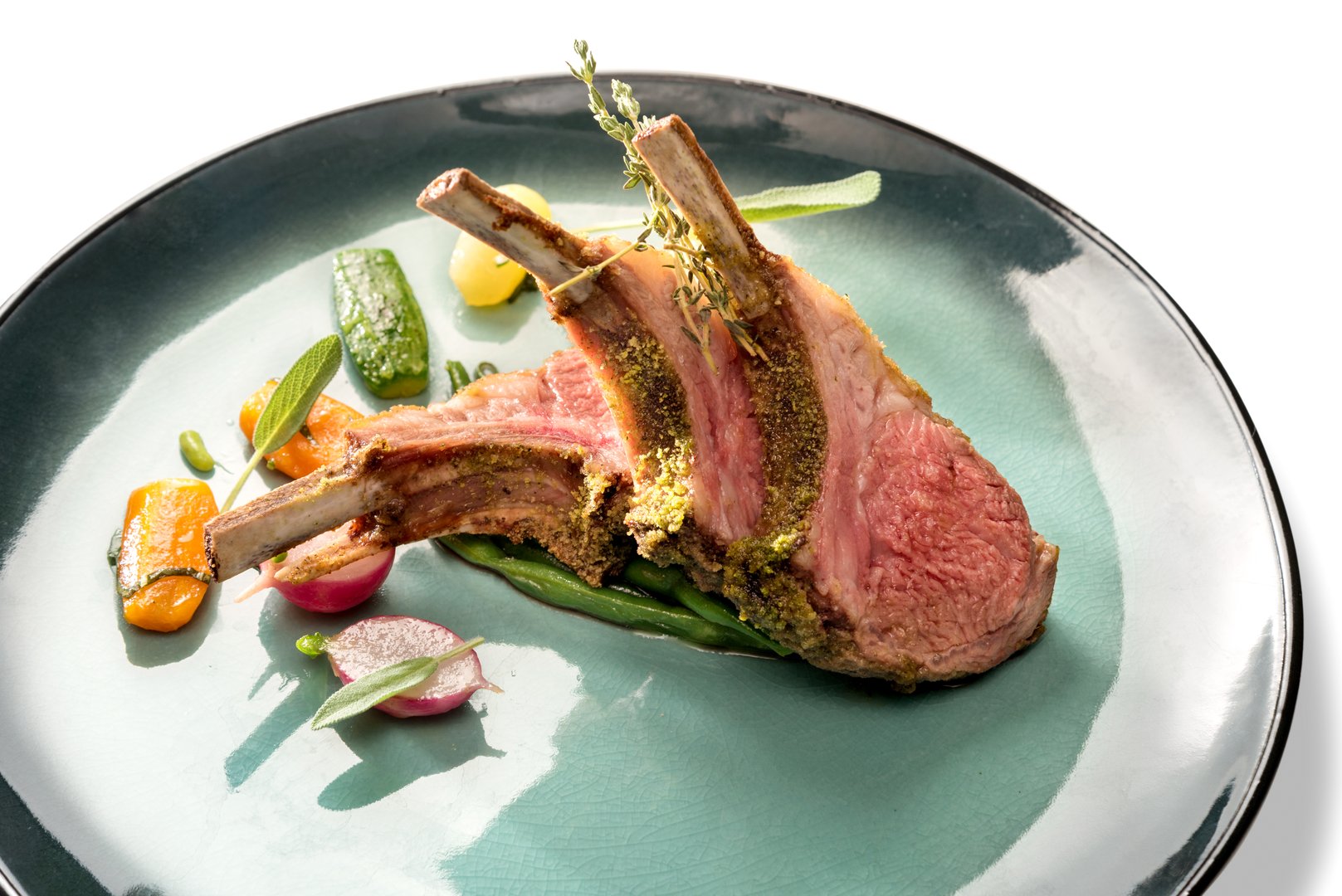 Crusted lamb ribs cutlets with bone in green dish with carrot, radishes and courgette, close up, isolated on white