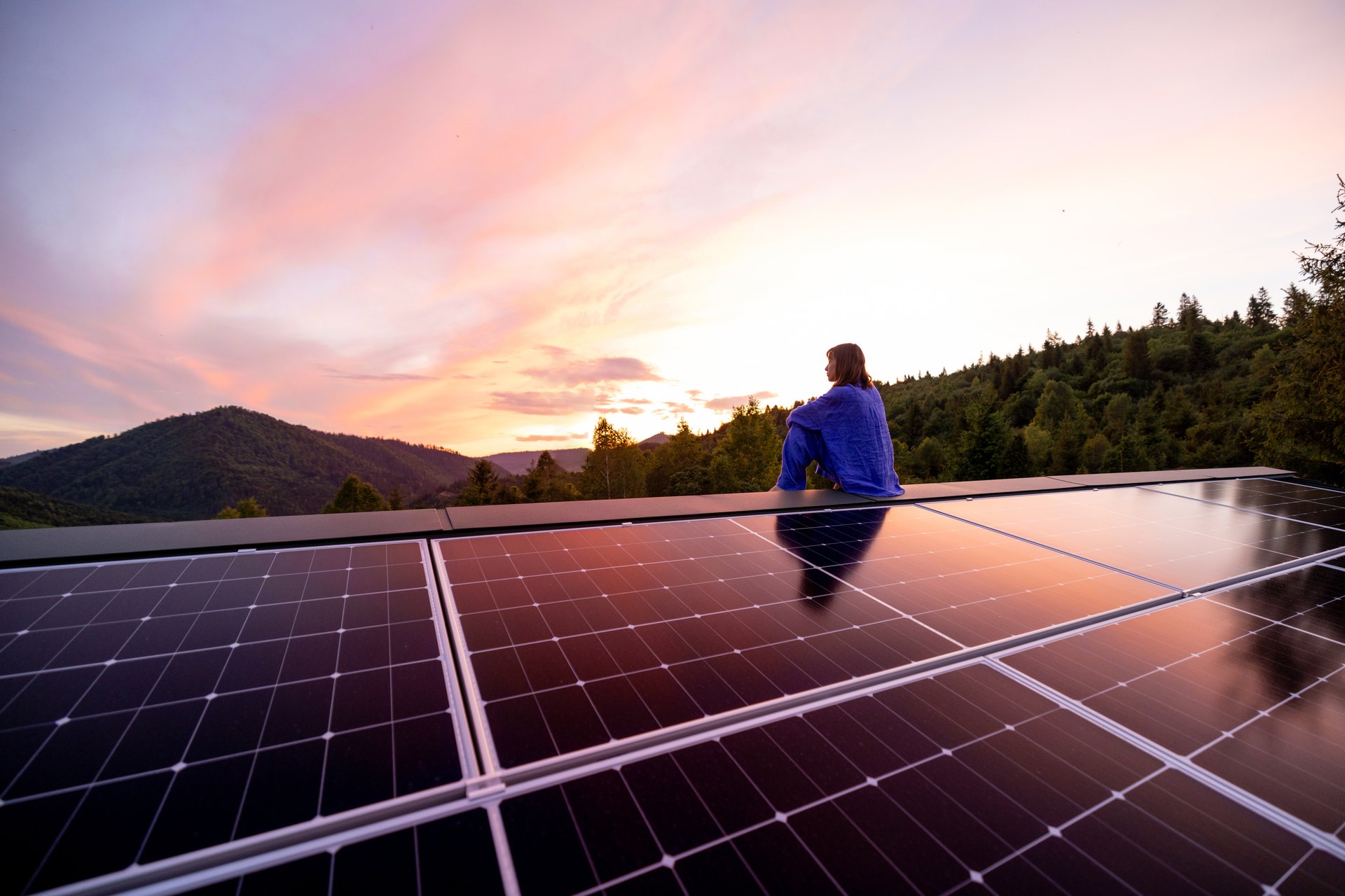 Rooftop with solar panels on house in mountains, woman sitting alone enjoying sunset. Energy independence, sustainability, self sufficient, and escapism to nature concept