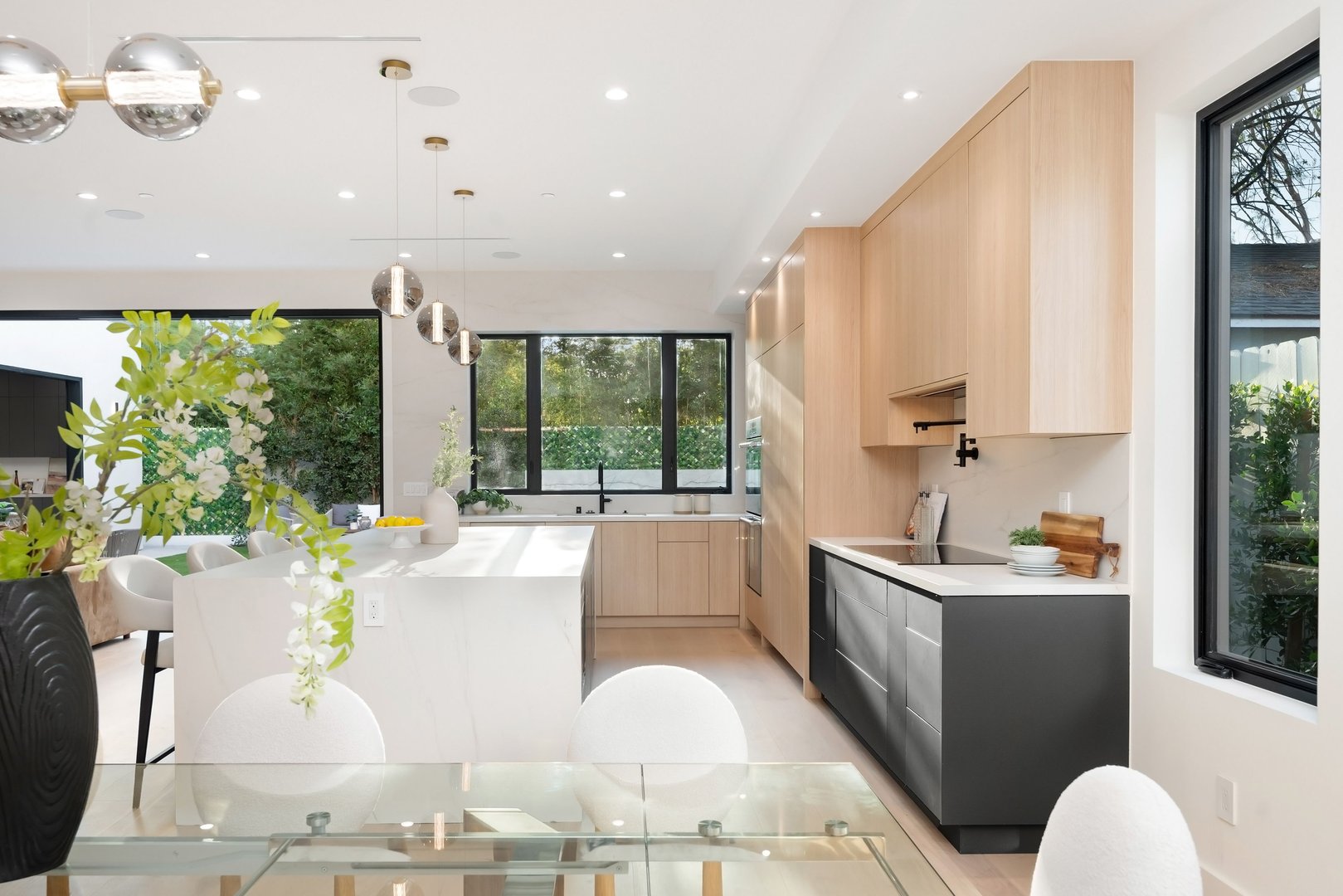 Modern kitchen interior with light wood cabinets, large windows, and stylish pendant lighting, featuring a sleek island and dining area.
