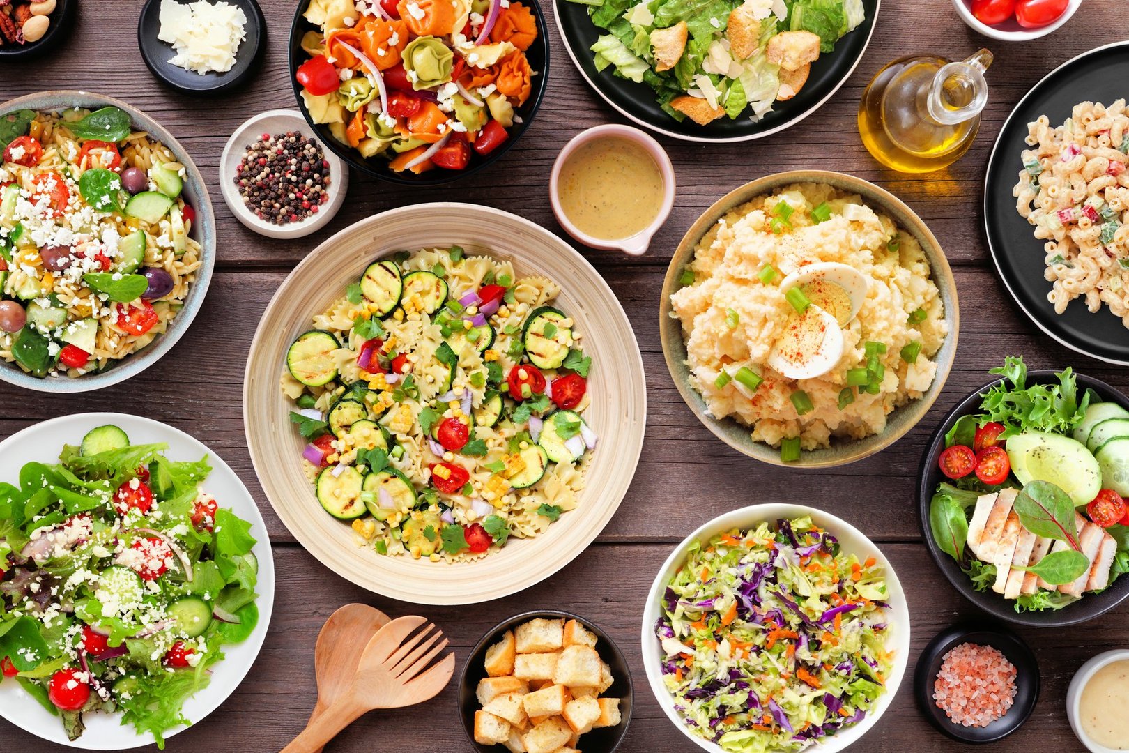 Assortment of delicious homemade salads. Top view table scene on a dark wood background. Summer food concept. Potato, caesar, pasta and green salads.