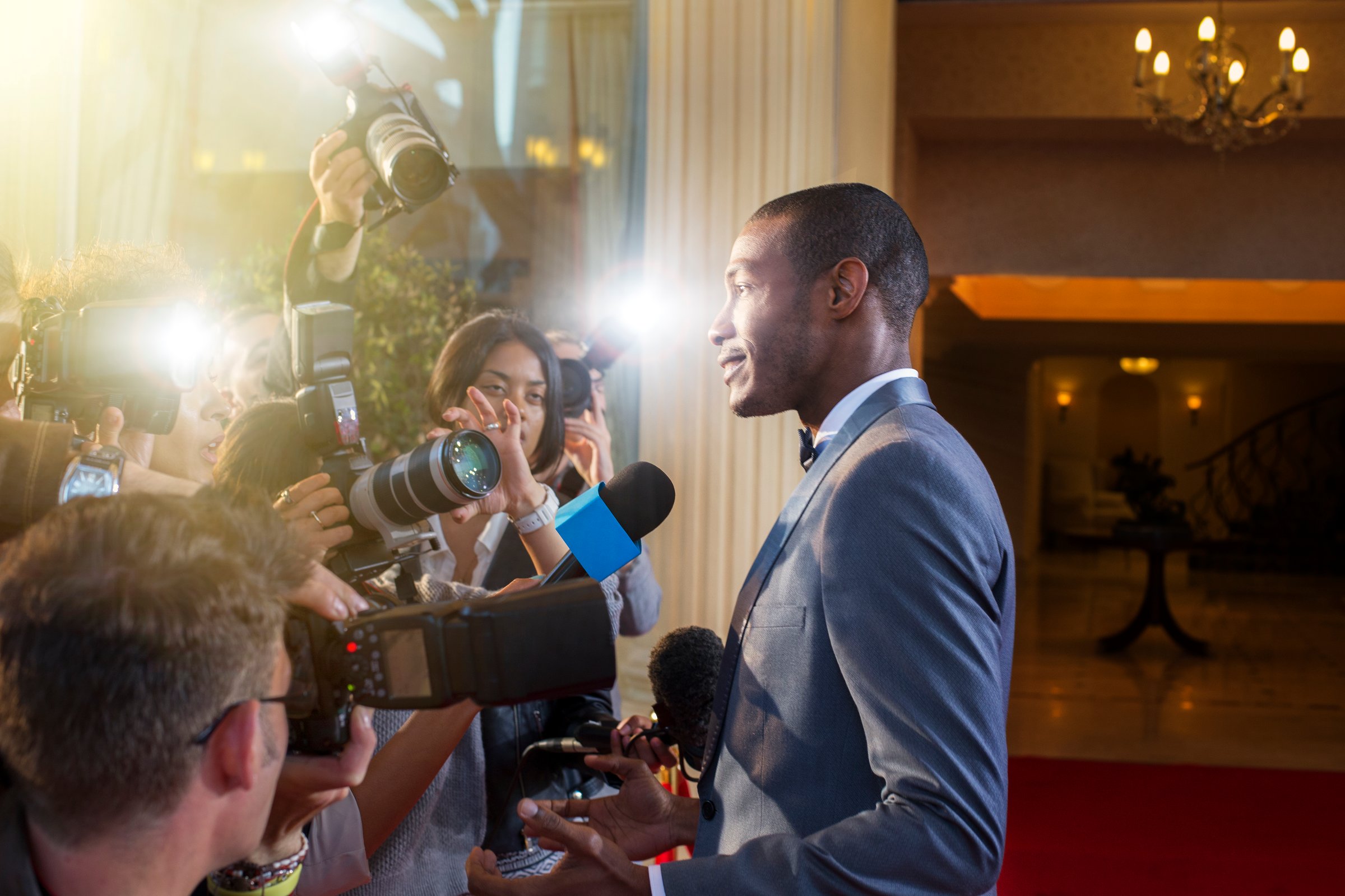 A man in a suit speaks to reporters and photographers on a red carpet, with bright camera flashes in a formal setting.