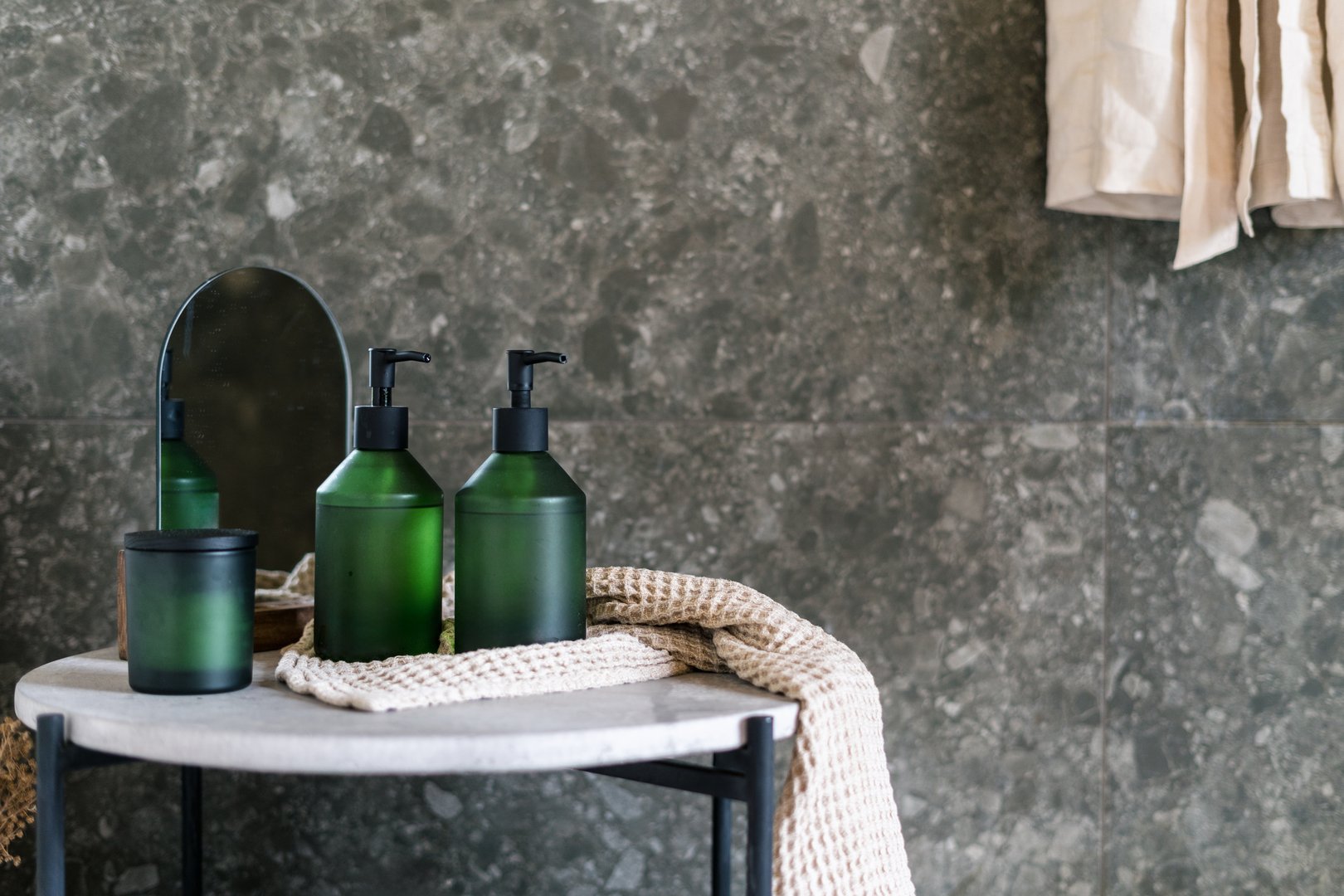 Three green soap dispenser bottles and a candle on a marble table in a modern bathroom. A terry towel drapes over the edge, enhancing the clean, minimalist decor in the hotel room