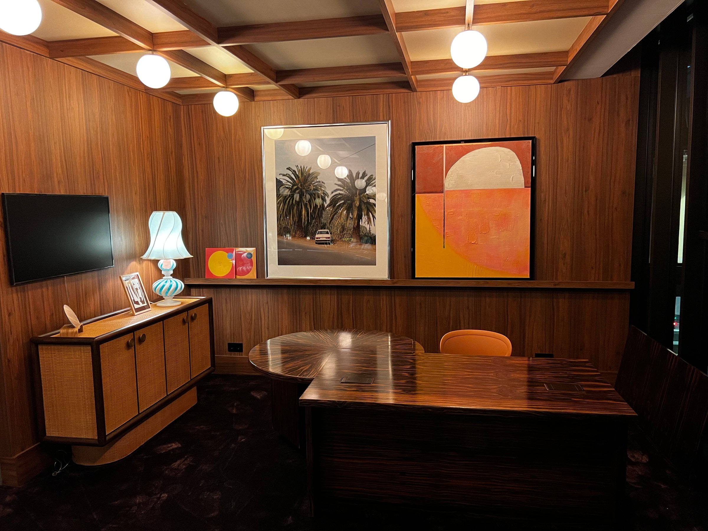 Retro-style office with wood-paneled walls, vintage furniture, modern art, and a warm lamp on a sideboard.