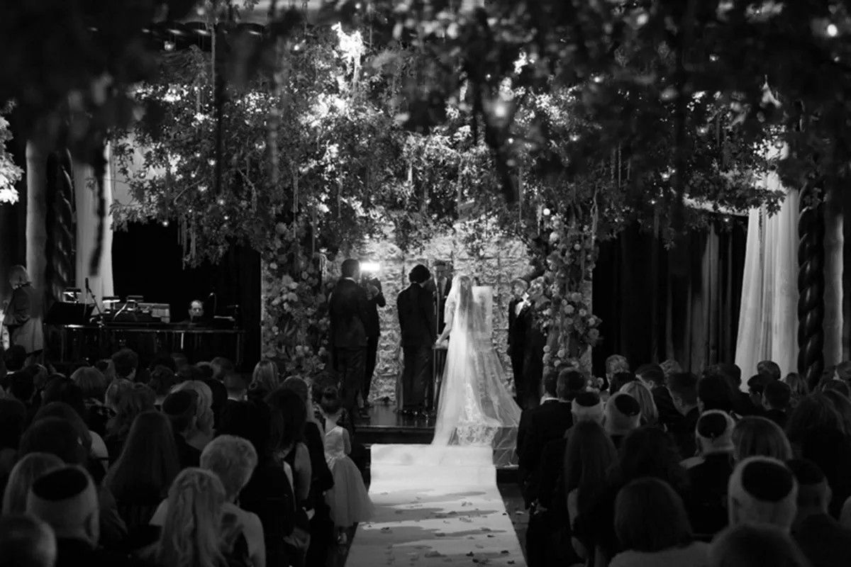 A black and white photo of a wedding ceremony with a bride and groom standing under a canopy, surrounded by guests.