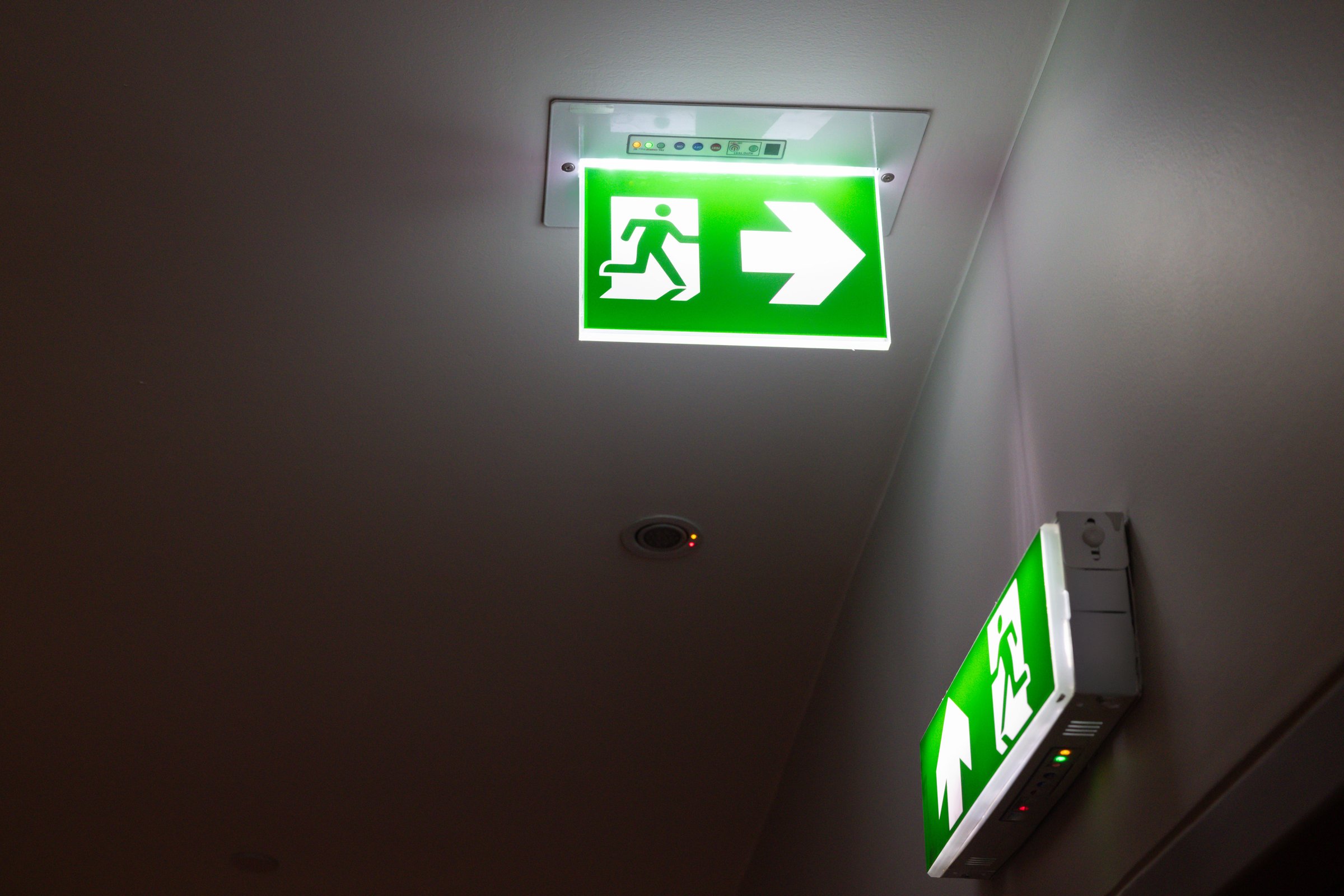 A green sign with a person running on it and an arrow pointing to the right. The sign is lit up and is hanging from the ceiling