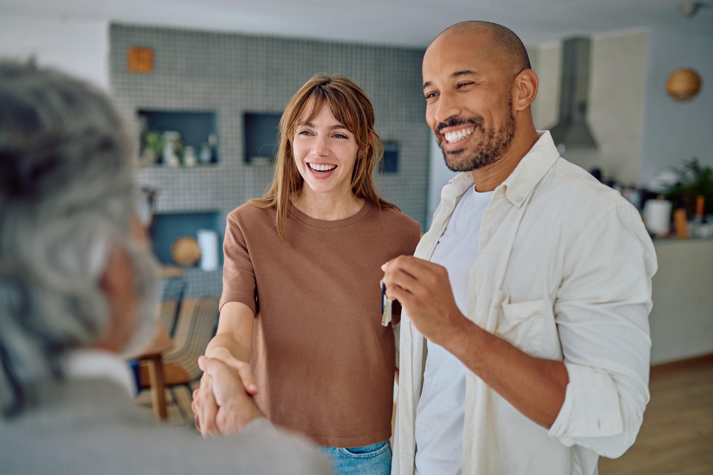 Young couple smiling while shaking hands with a real estate agent, joyfully receiving the keys to their new home after finalizing the purchase