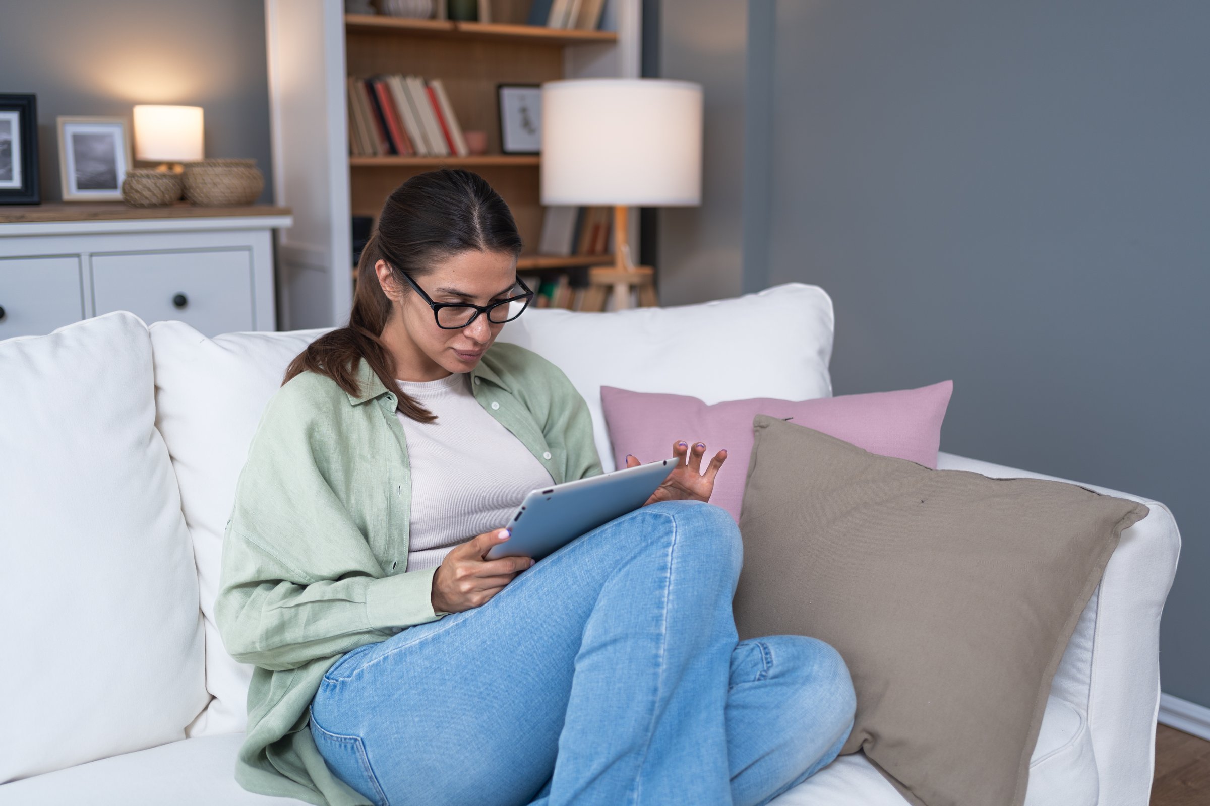 Relaxed young adult caucasian woman read a book on modern device reader comfortably sitting on the couch at home Portrait of pretty female people wearing eyeglasses and using digital tablet connection