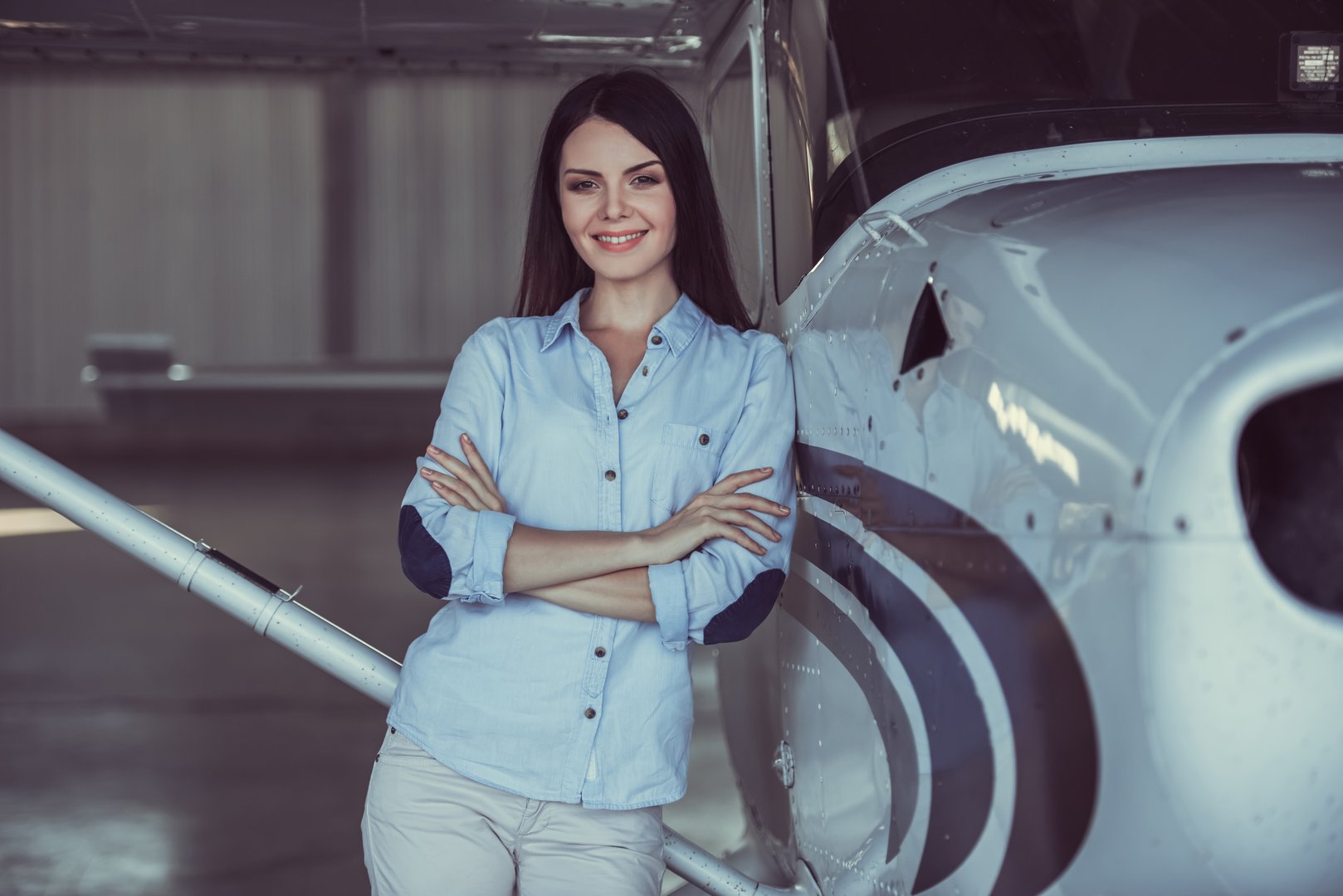 Beautiful woman is looking at camera and smiling while standing with crossed arms near the aircraft in hangar