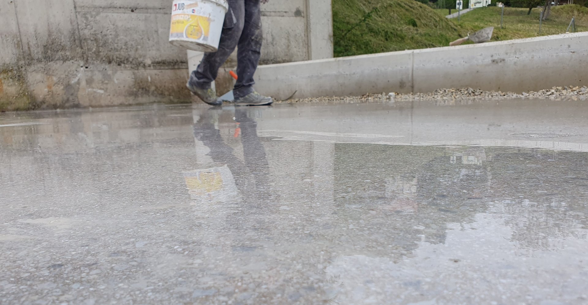 Person carrying a paint bucket walking on a wet concrete surface, with reflections visible on the ground.