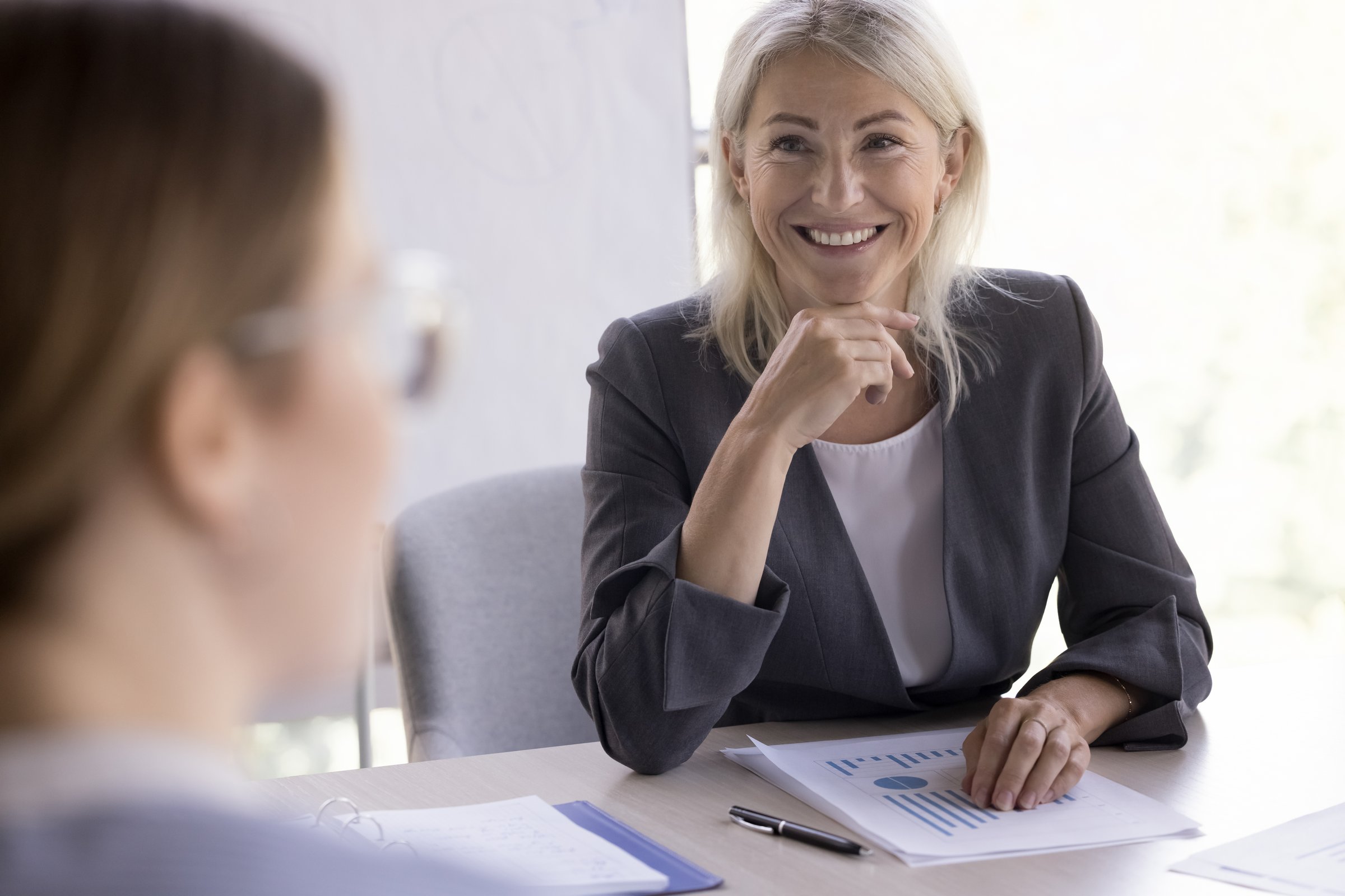 Cheerful confident mature businesswoman talking to partners on negotiation meeting. Boss, business leader woman speaking to young employee, smiling. Mentor giving lesson, lecture to interns