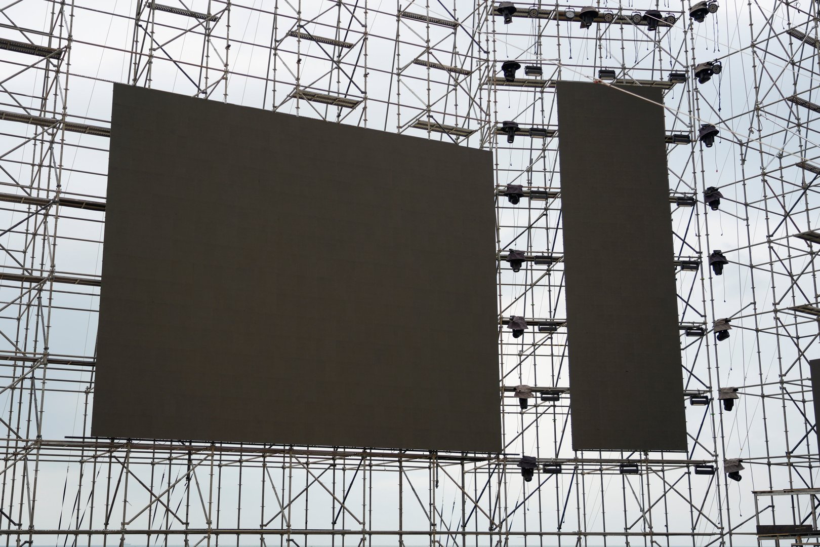 A massive stage is being constructed with scaffolding, featuring large blank screens ready for visuals. The activity occurs in an outdoor setting under a cloudy sky in daytime.
