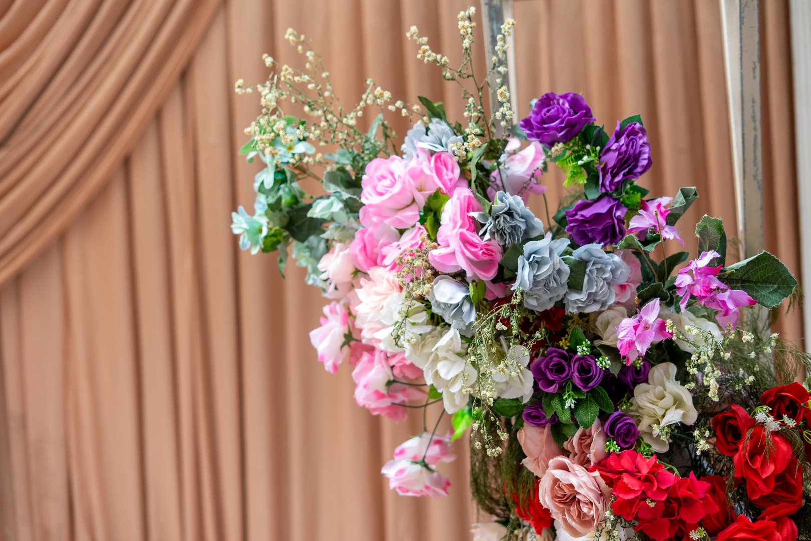 A close-up shot of a lavish artificial flower arrangement featuring pink, purple, red, white, and light blue blooms, set against a soft, blurred beige curtain background. Ideal for celebration decor.